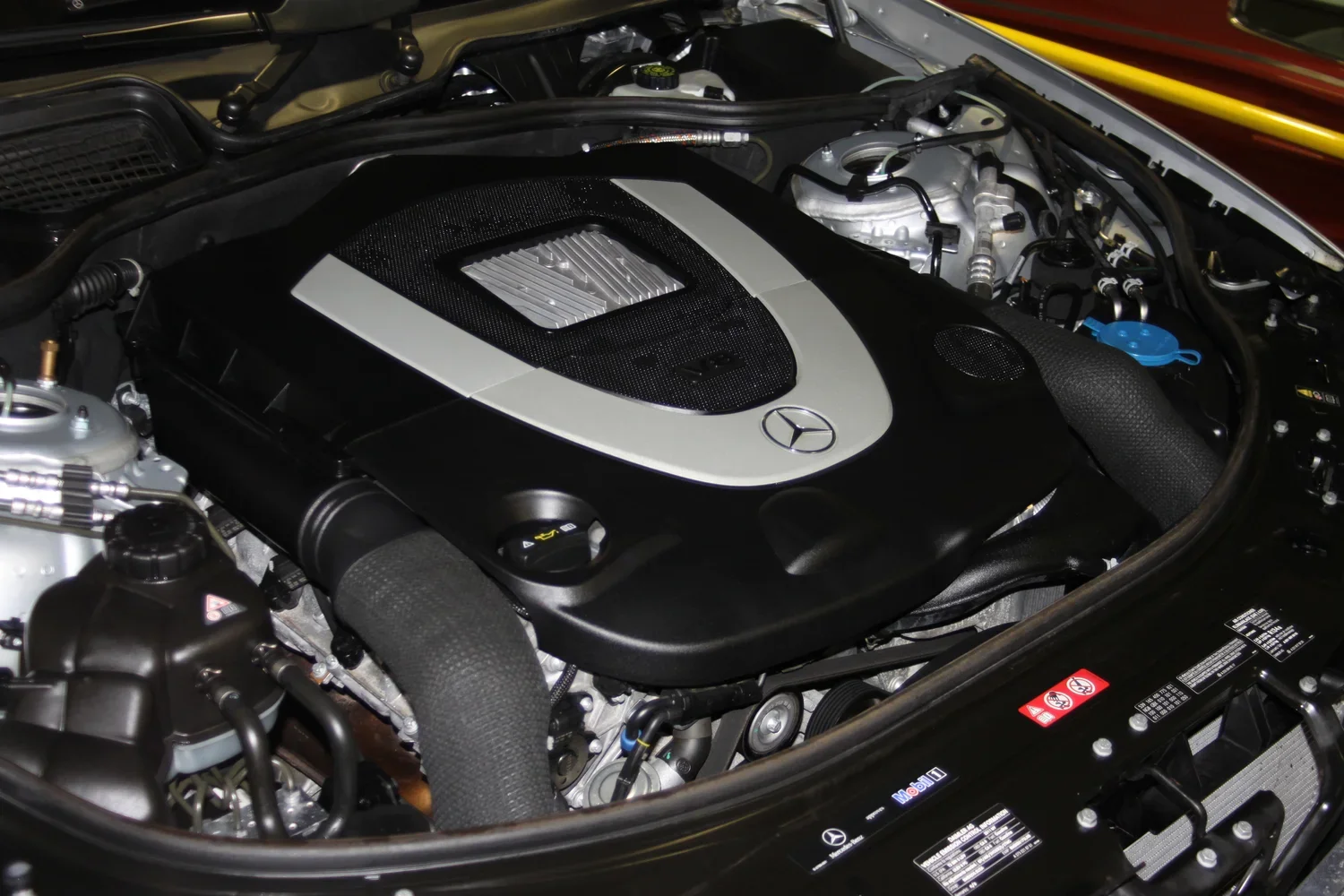 Close-up of a Mercedes-Benz engine bay showing the black engine cover with Mercedes logo and silver accents, surrounded by various engine components and hoses.