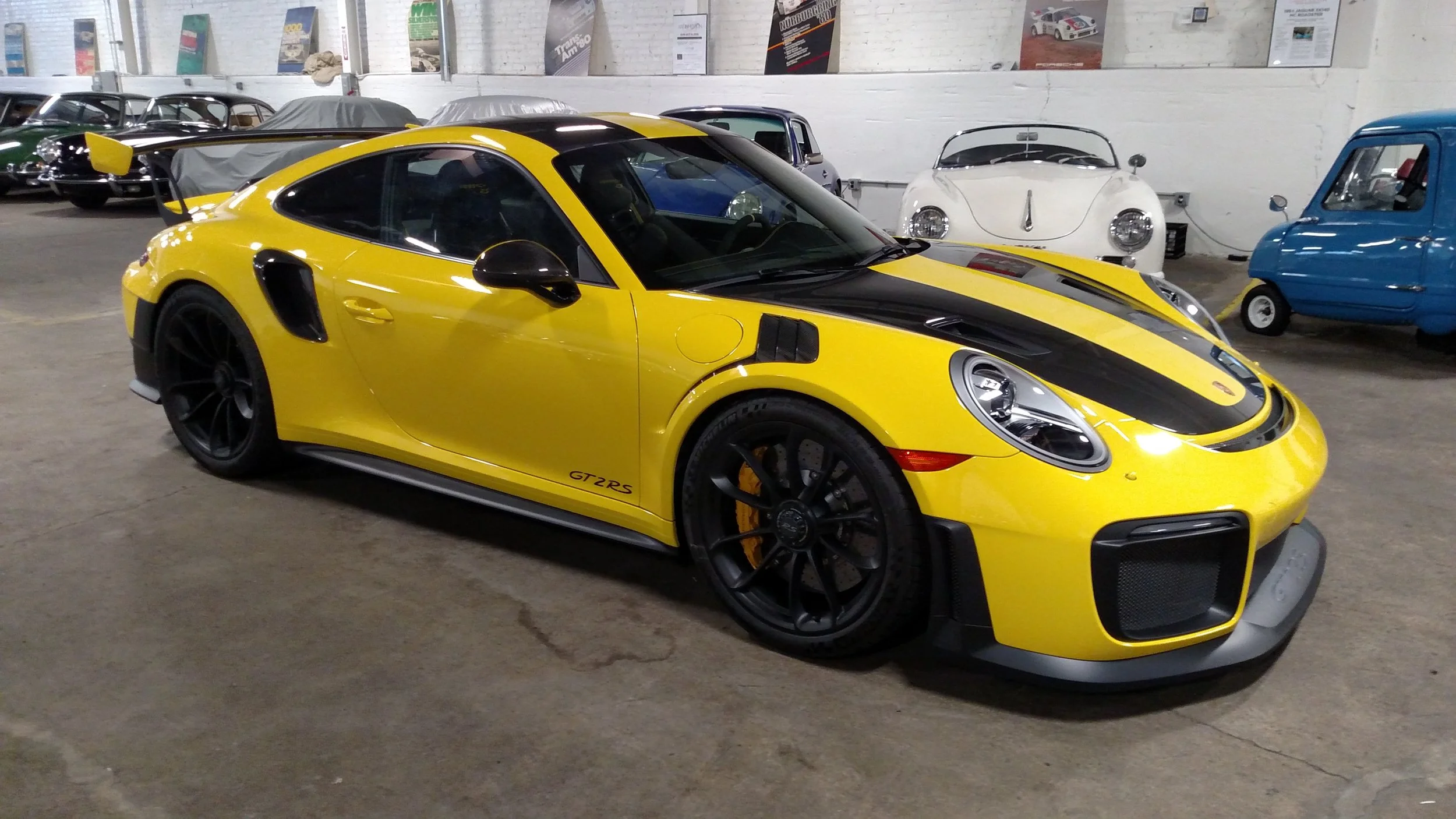 Yellow Porsche 911 GT2 RS with black accents, black wheels, and a large rear wing, parked inside a museum with other vintage cars in the background.