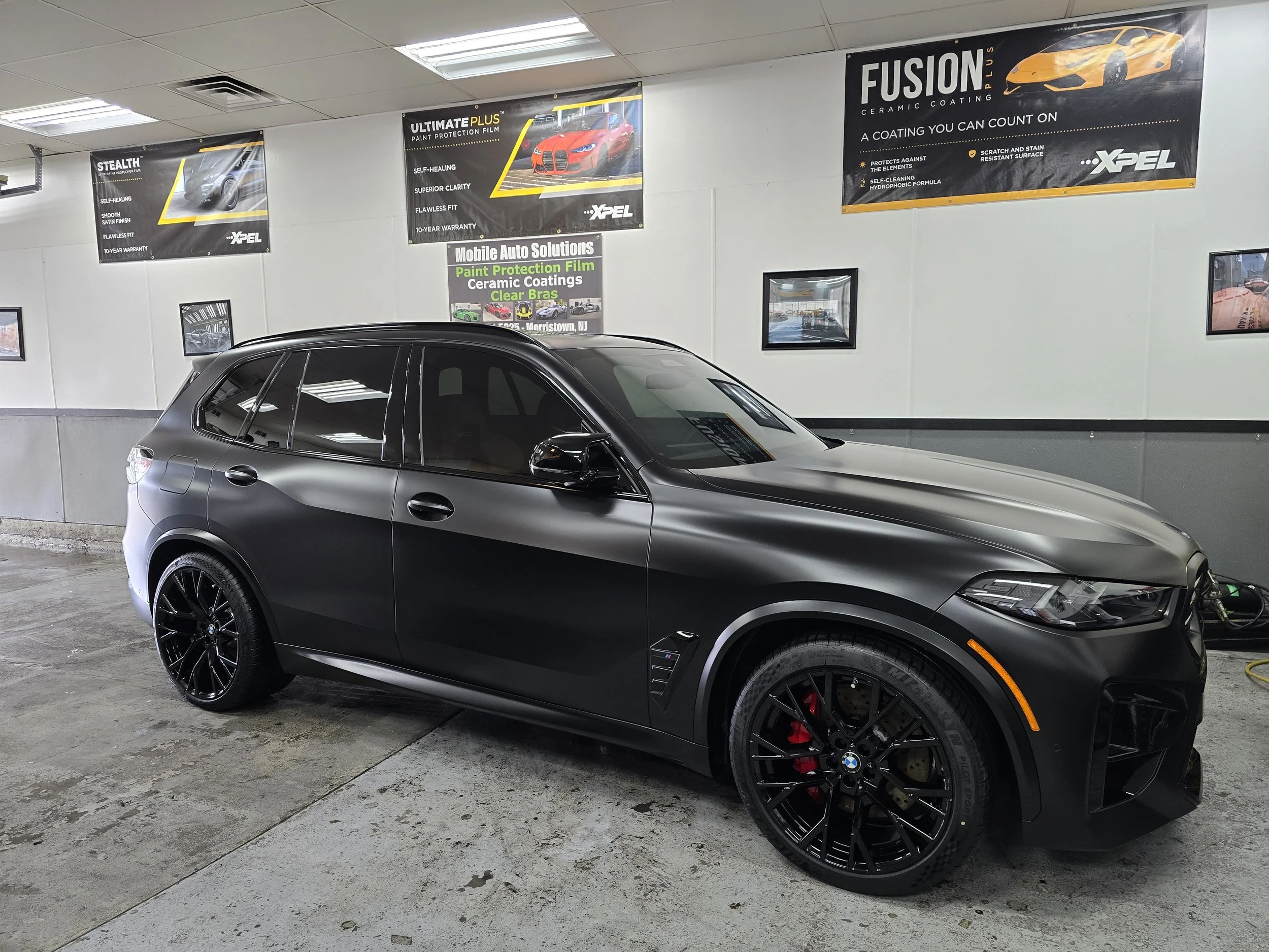 Black BMW SUV parked inside a garage or service area with promotional posters on the wall behind it, including advertisements for car paint protection and ceramic coating.