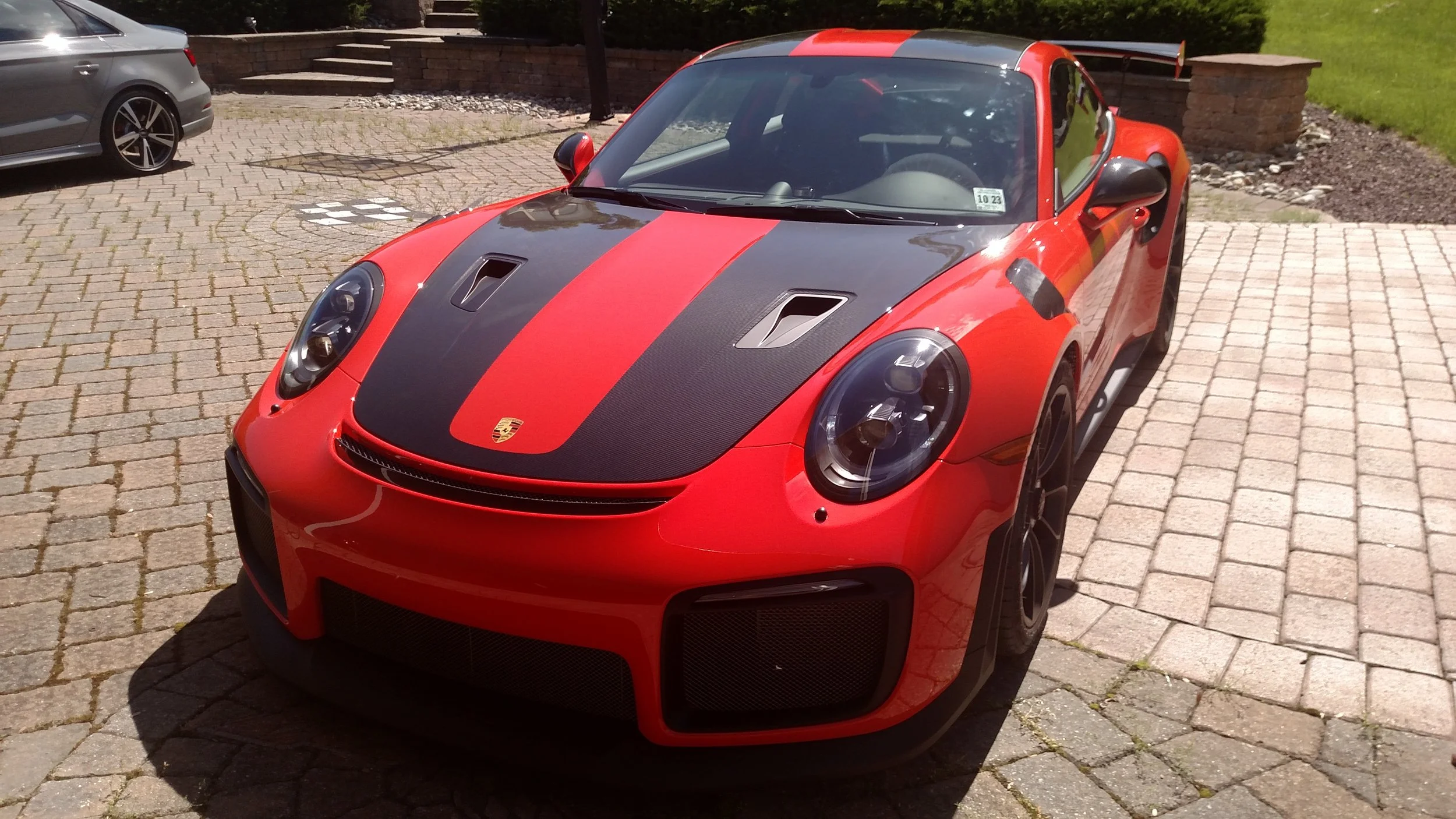Red Porsche sports car with black racing stripe and carbon fiber accents parked on a brick driveway.