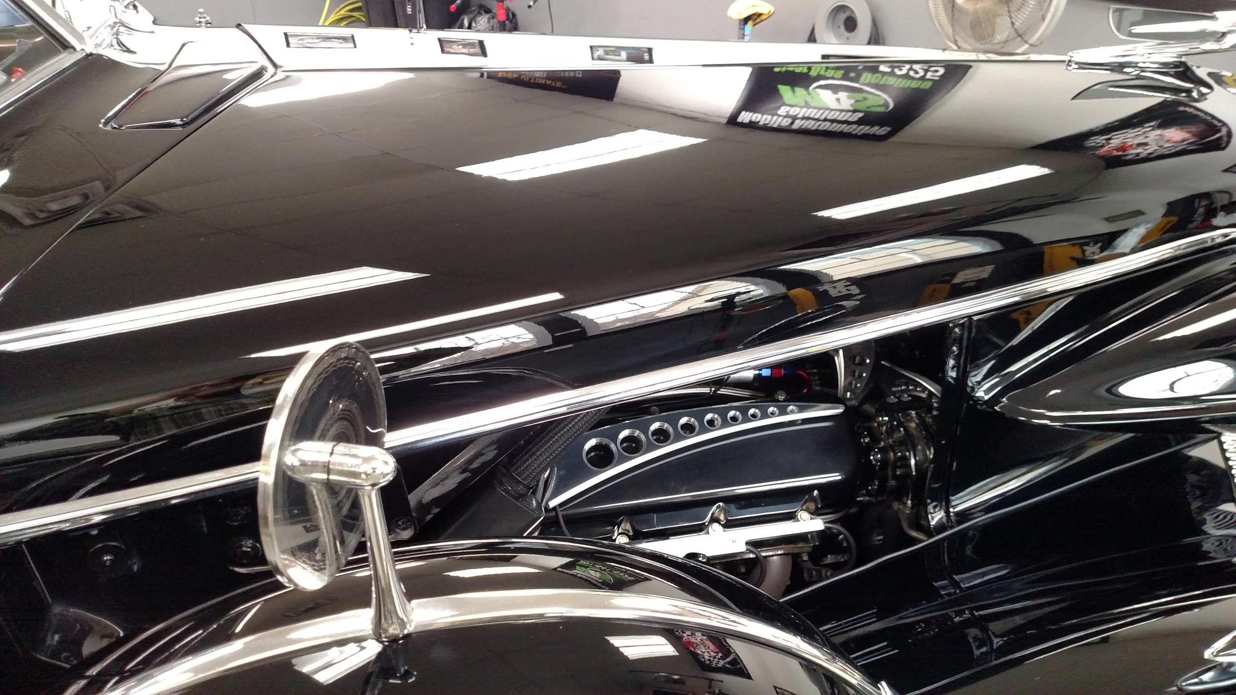 Close-up of a classic black vintage race car interior with a steering wheel, dashboard, and parts of the exterior showing a mirror and reflections.