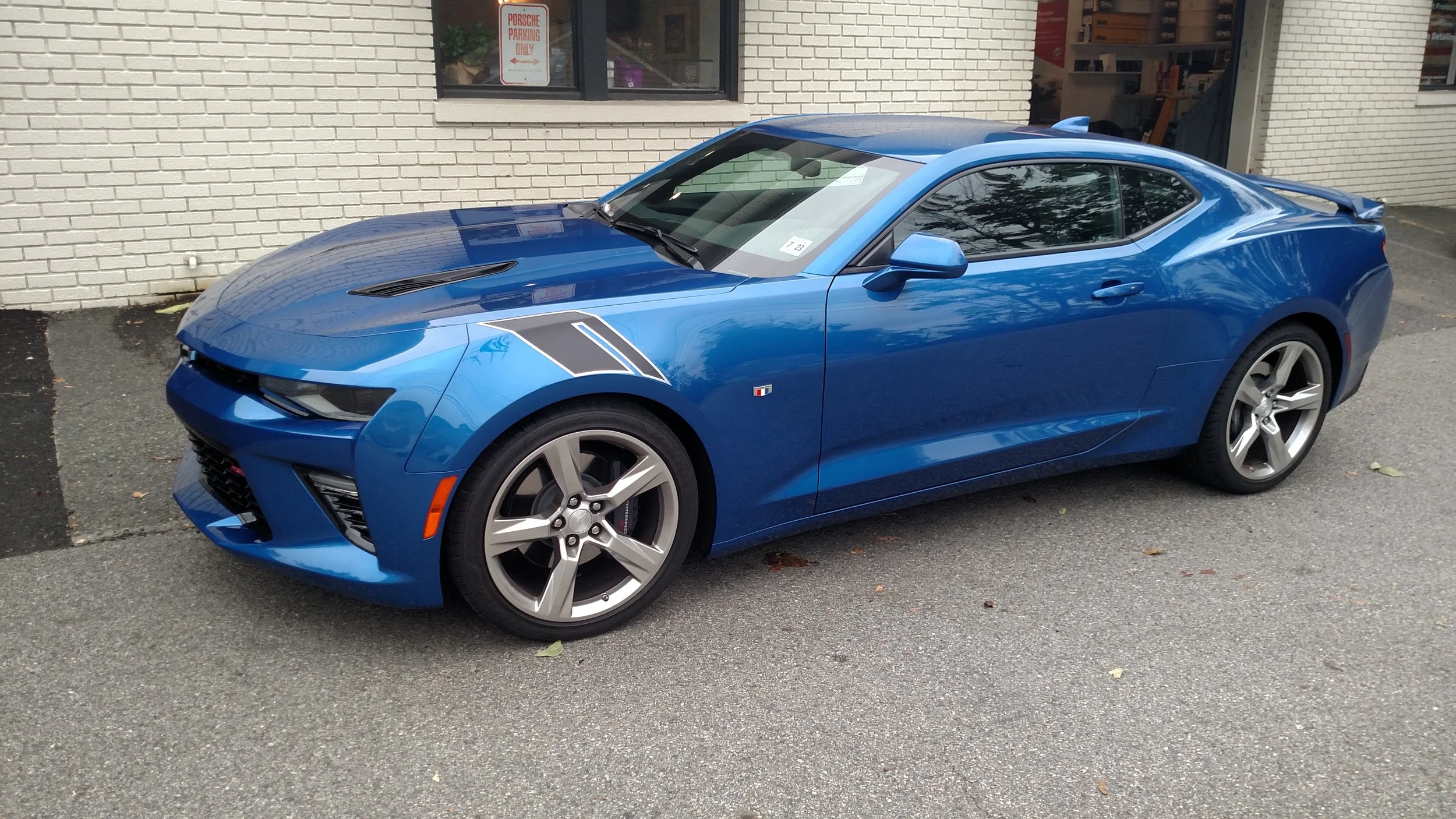 Blue sports car parked outside near a white brick building with a window and a 'Porsche Parking Only' sign.