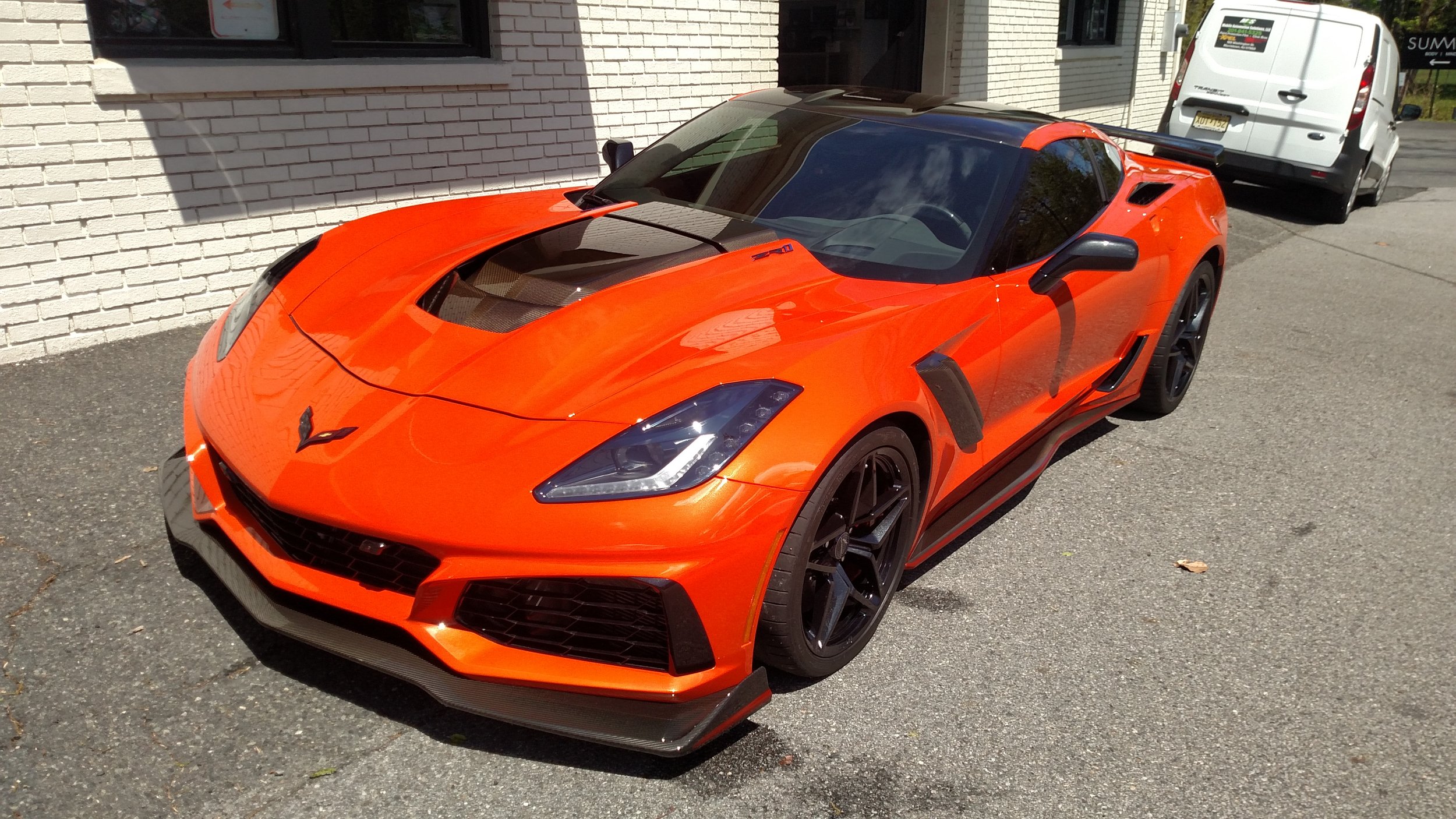Orange Chevrolet Corvette sports car parked on a street near a white brick building with a white van and another vehicle in the background.