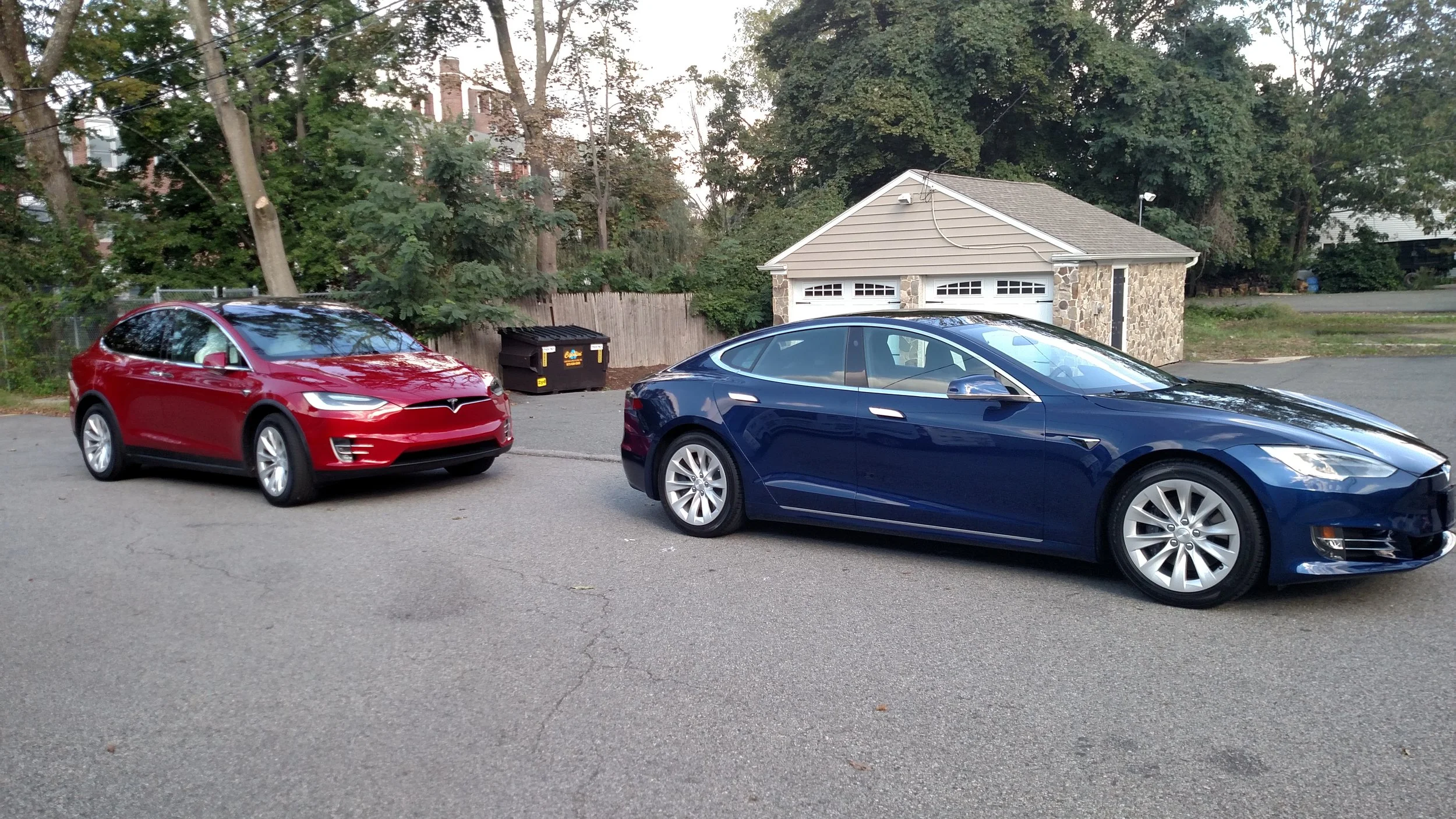 A red Tesla Model S and a blue Tesla Model S parked on a residential street with a stone garage, trees, and a fence in the background.