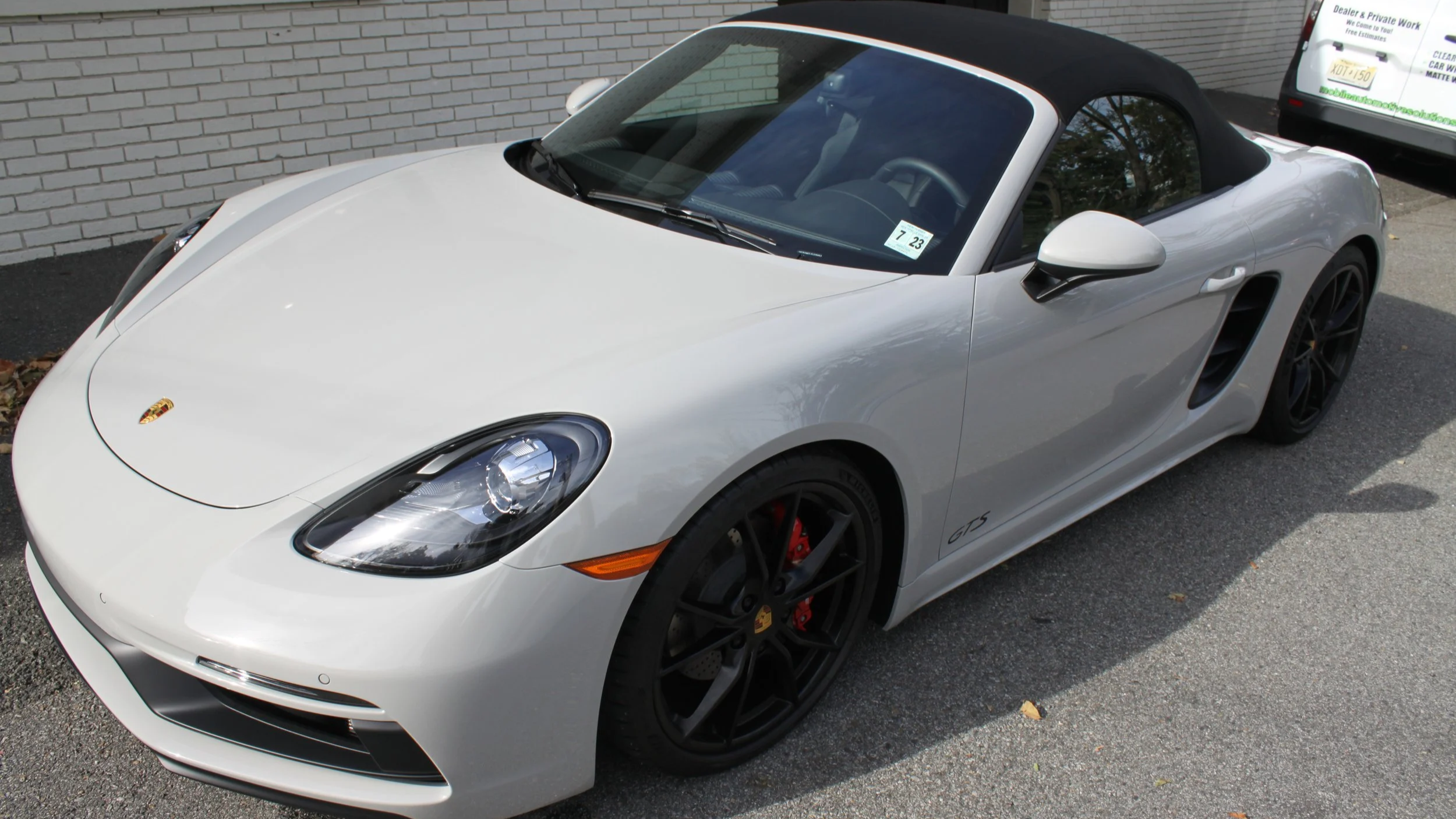 A white Porsche 718 Boxster GTS with a black convertible top parked on asphalt, with another vehicle partially visible in the background.