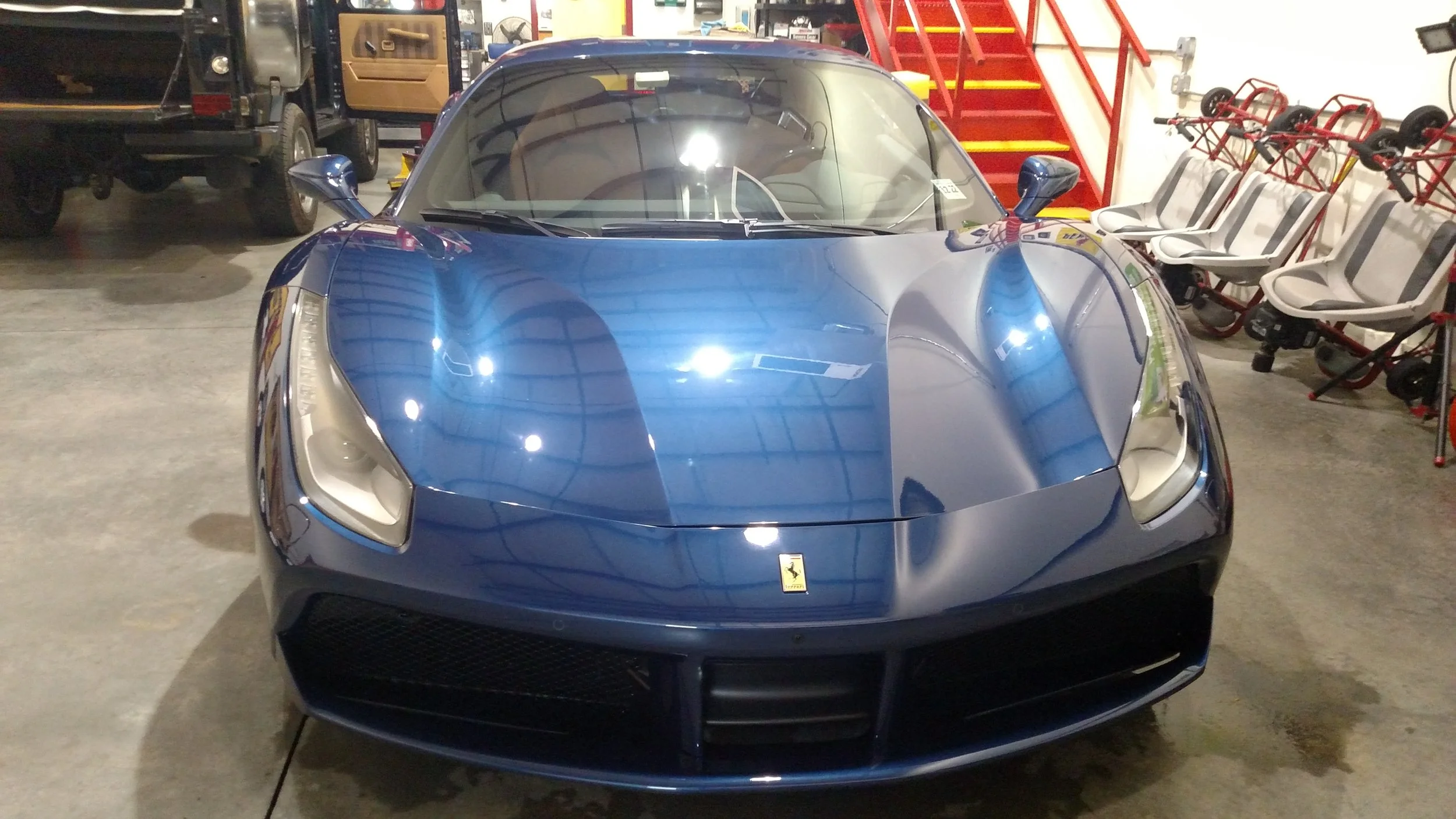 Front view of a blue Ferrari sports car inside a garage or showroom, with chairs and red stairs in the background.