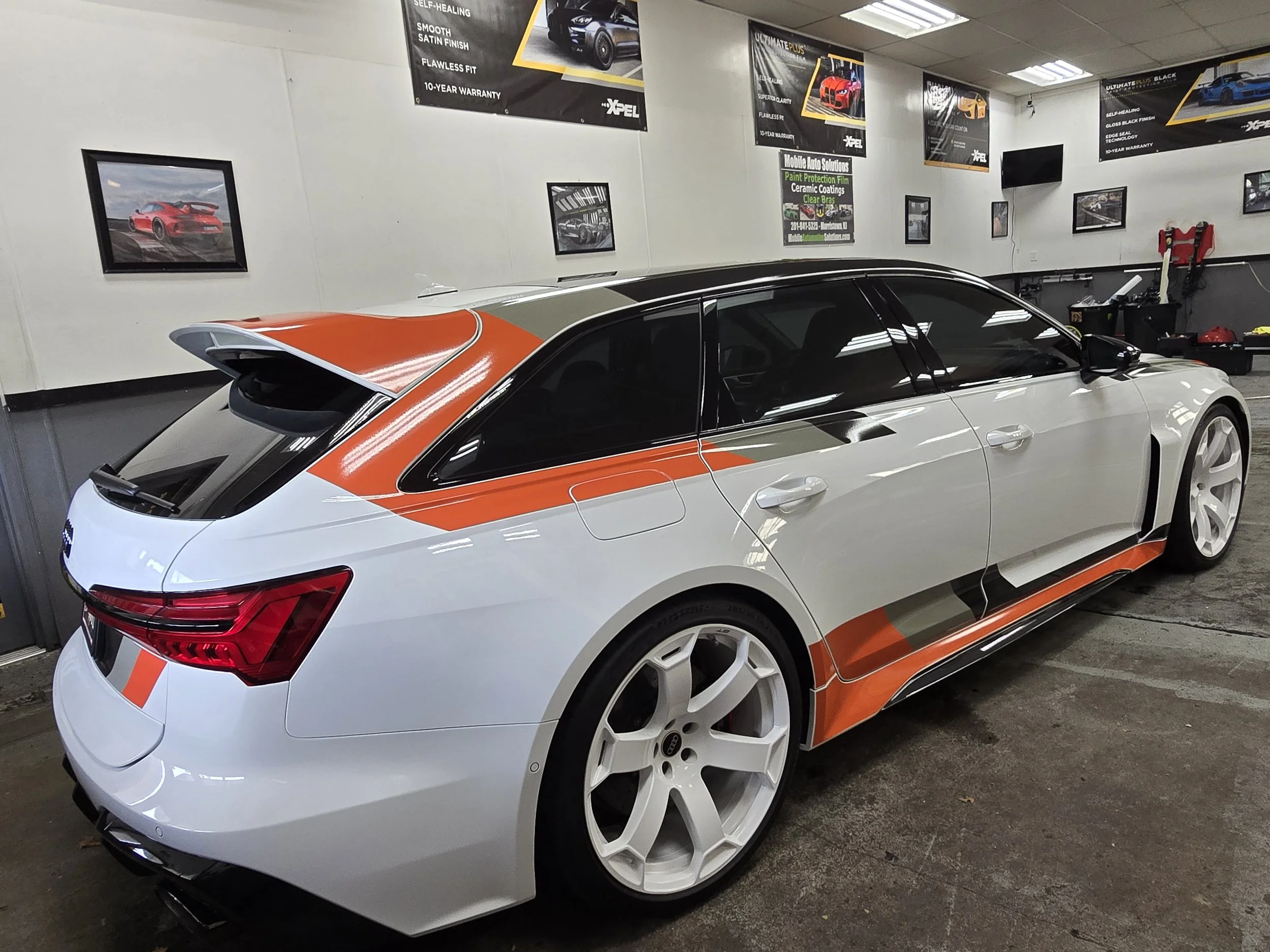 White sports car with orange and black accents parked inside a garage with car posters and advertisements on the walls.