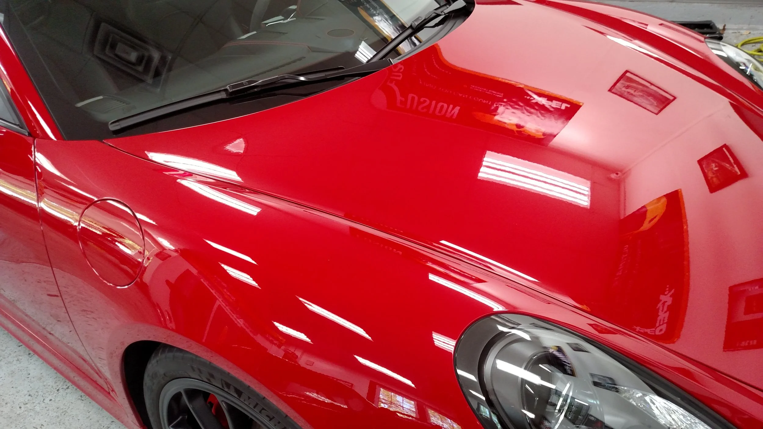 Close-up of a shiny red sports car with a black hood, showing reflections of ceiling lights and framed pictures on the smooth surface.