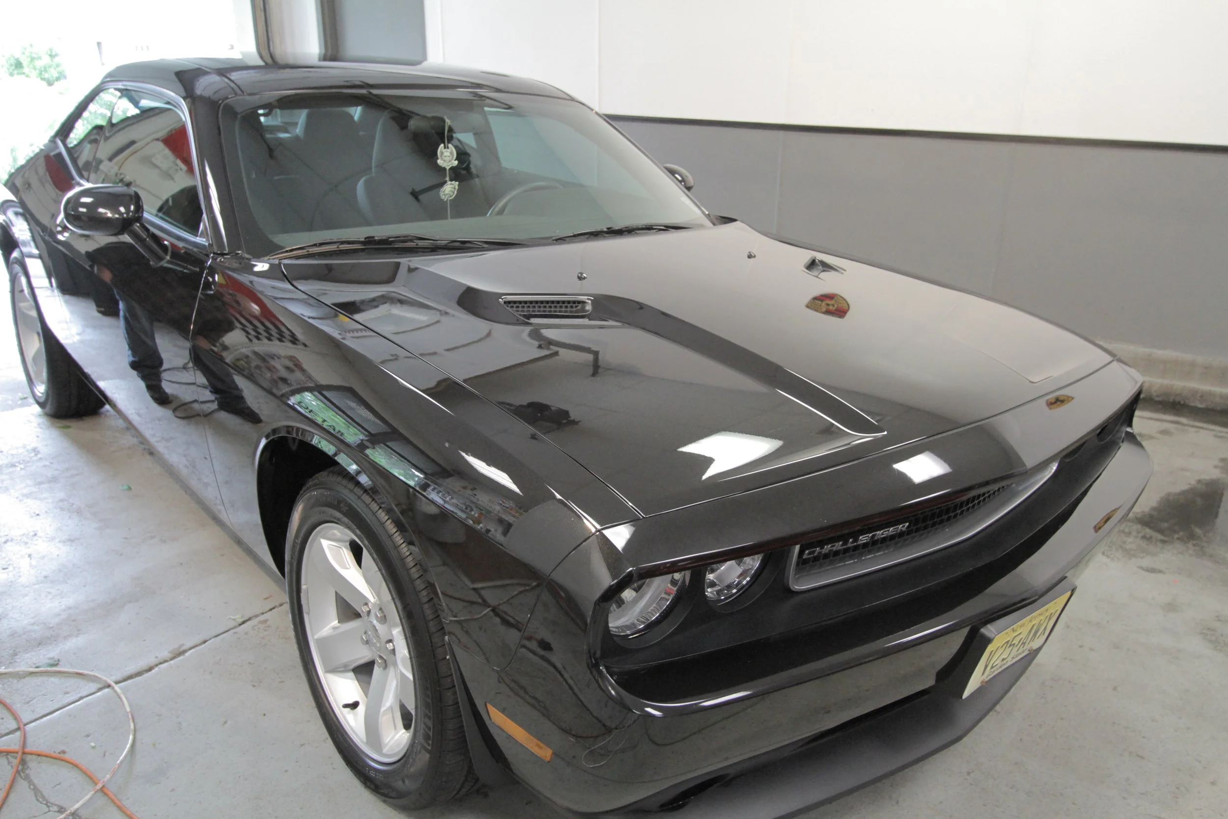 A black Dodge Challenger sports car parked inside a garage, with reflections of the surroundings visible on its shiny surface.