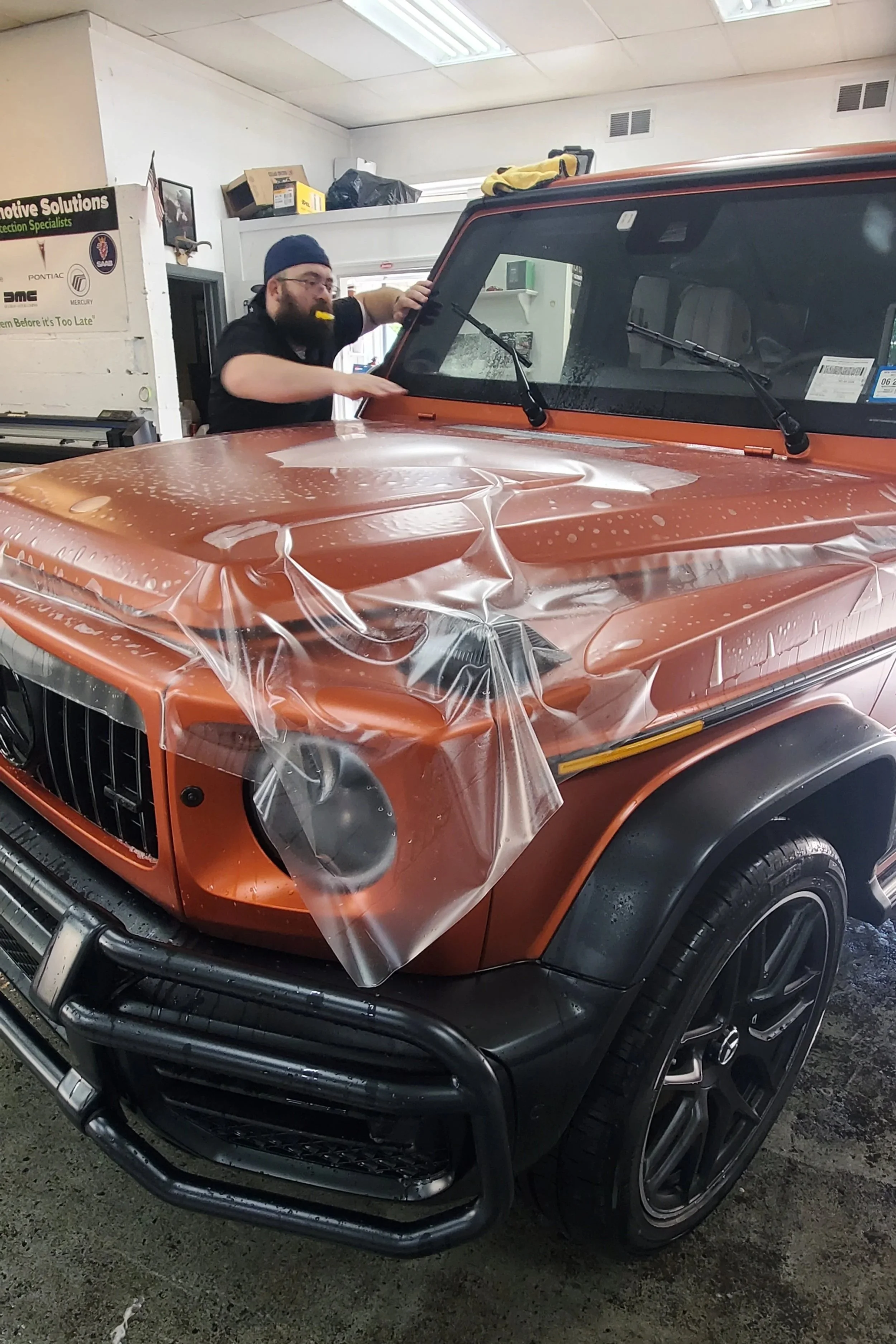A person is applying a protective film or wrapping to a metallic orange Jeep vehicle inside a workshop or garage. XPEL Stealth