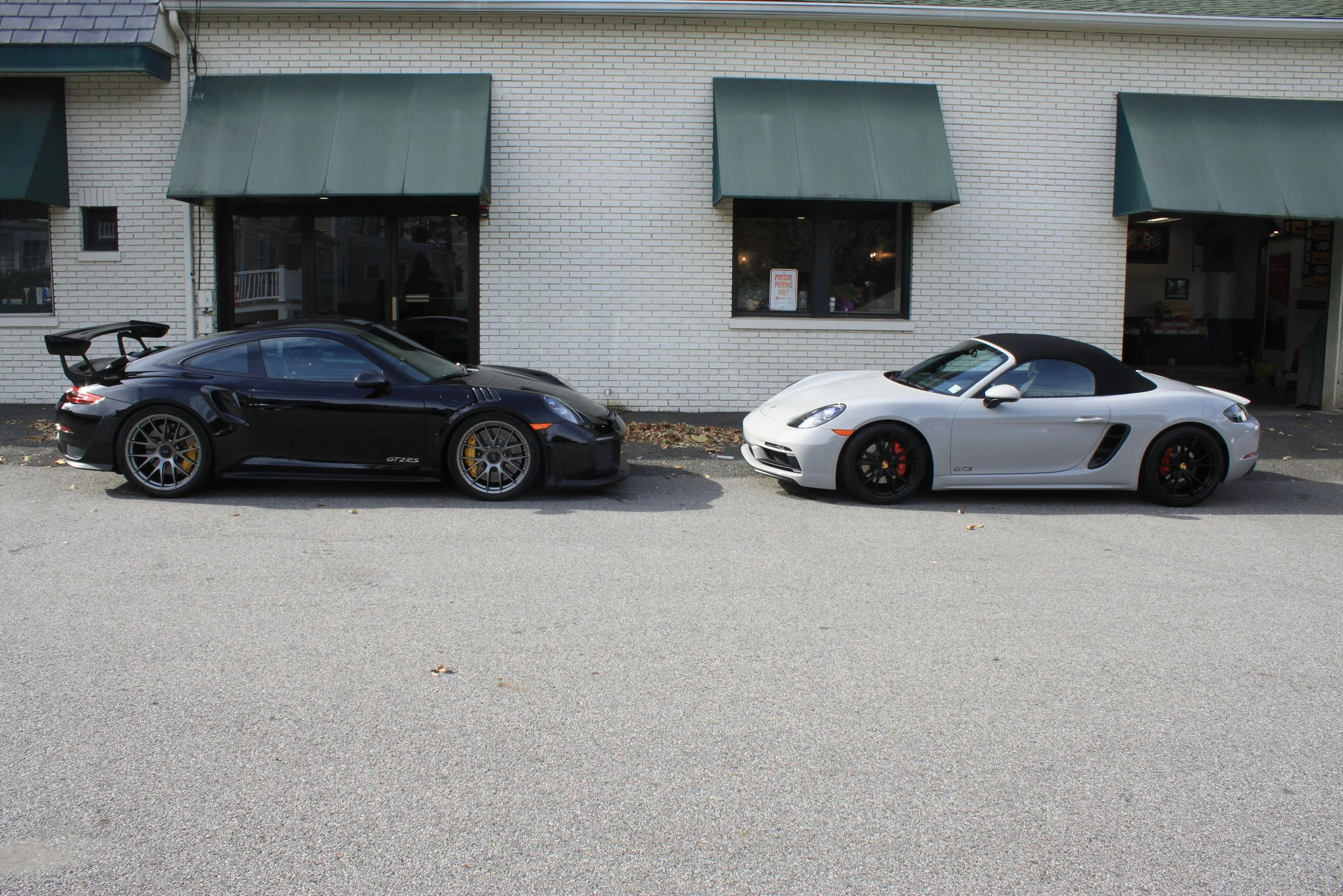 Two Porsche sports cars, one black and one white, parked on the street in front of a building with white brick walls and green awnings.
