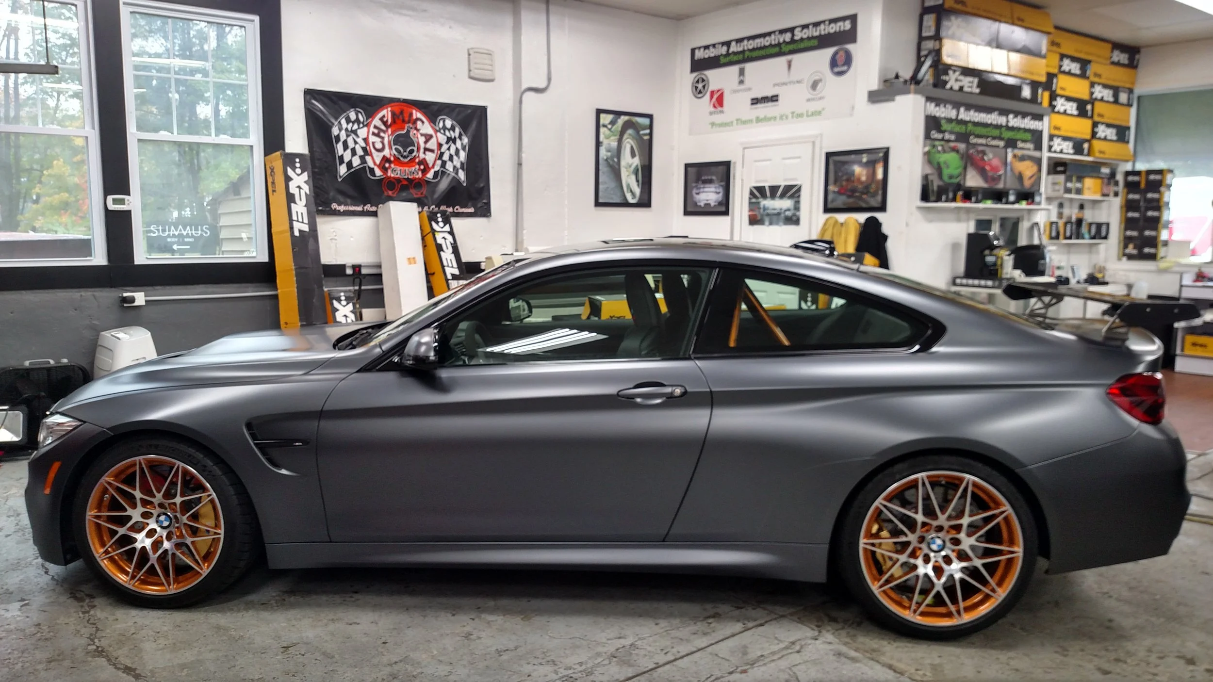 A silver BMW coupe with orange rims parked inside a garage, surrounded by automotive posters and tools.