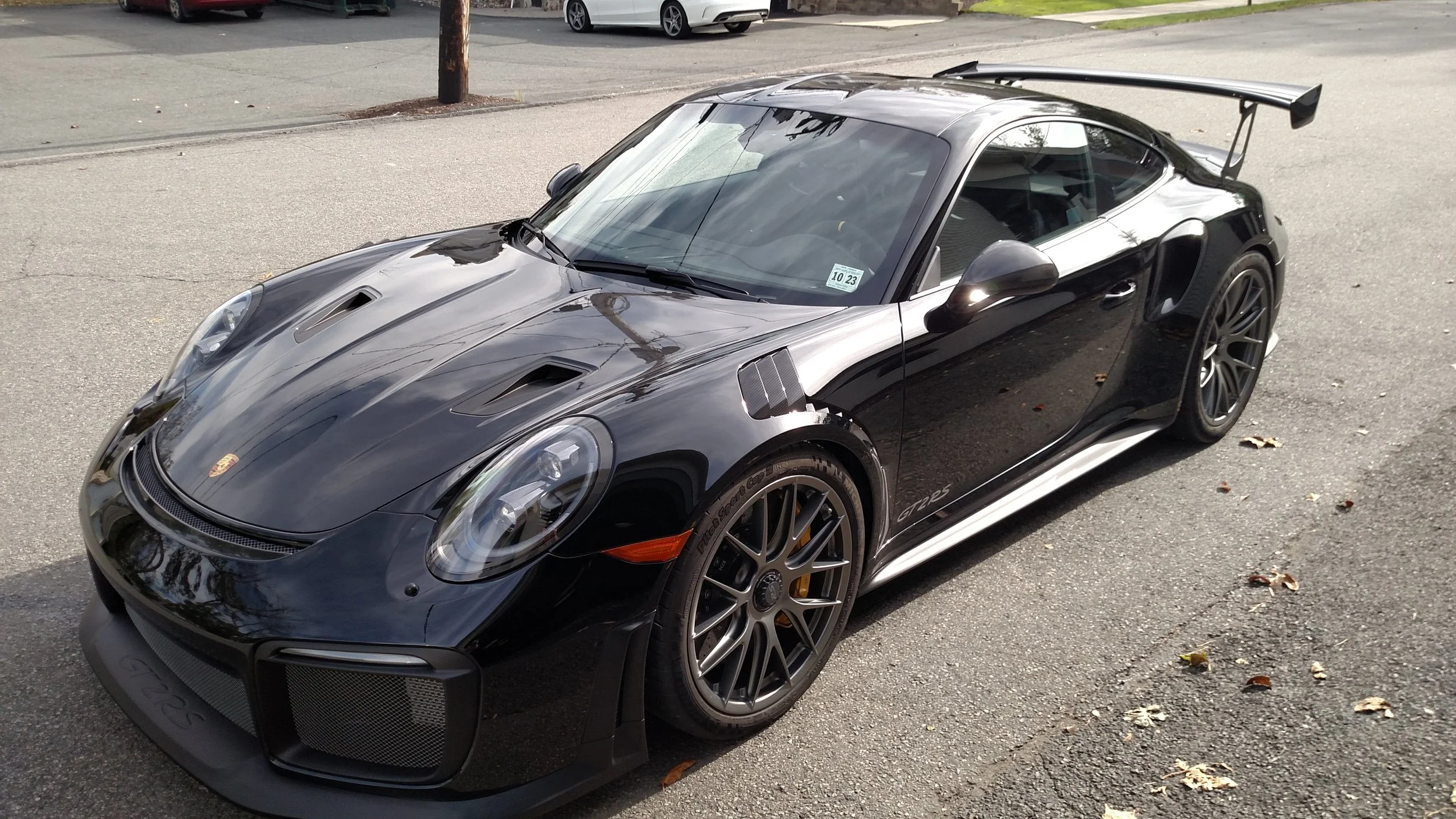 Black Porsche 911 GT2 RS parked on the street with a large rear wing, front vents, and racing tires, reflecting the surrounding sky and trees.