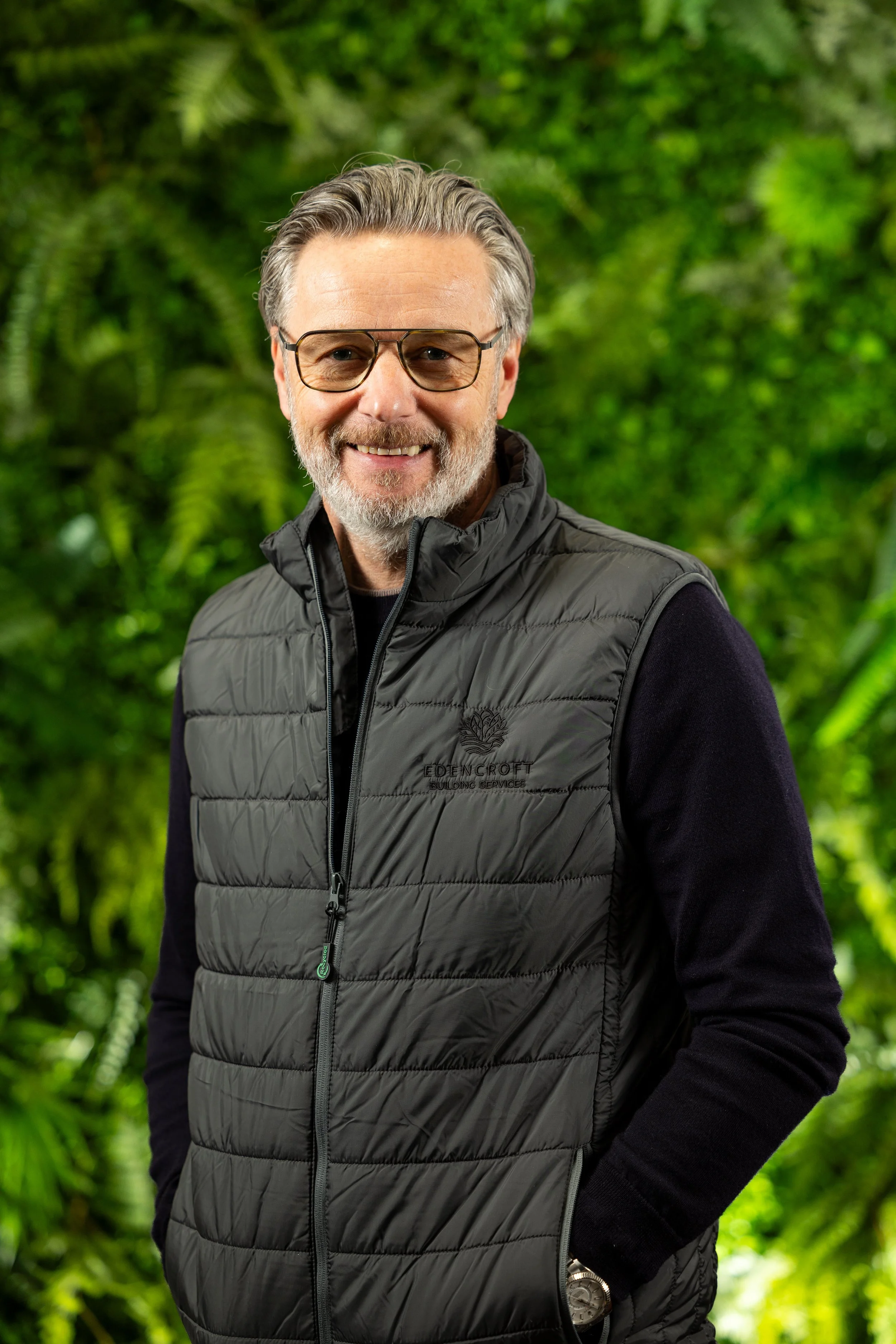 A middle-aged man with gray hair and beard, wearing glasses and a light blue button-up shirt with a small logo, smiling in front of a black-and-white cityscape photograph.