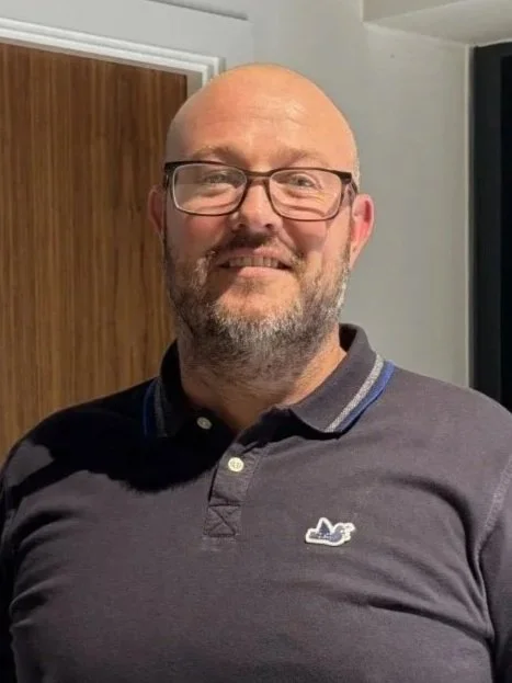A smiling middle-aged man with gray hair, beard, and glasses, wearing a pink dress shirt and dark blazer, standing in front of a wall with multiple framed photographs.