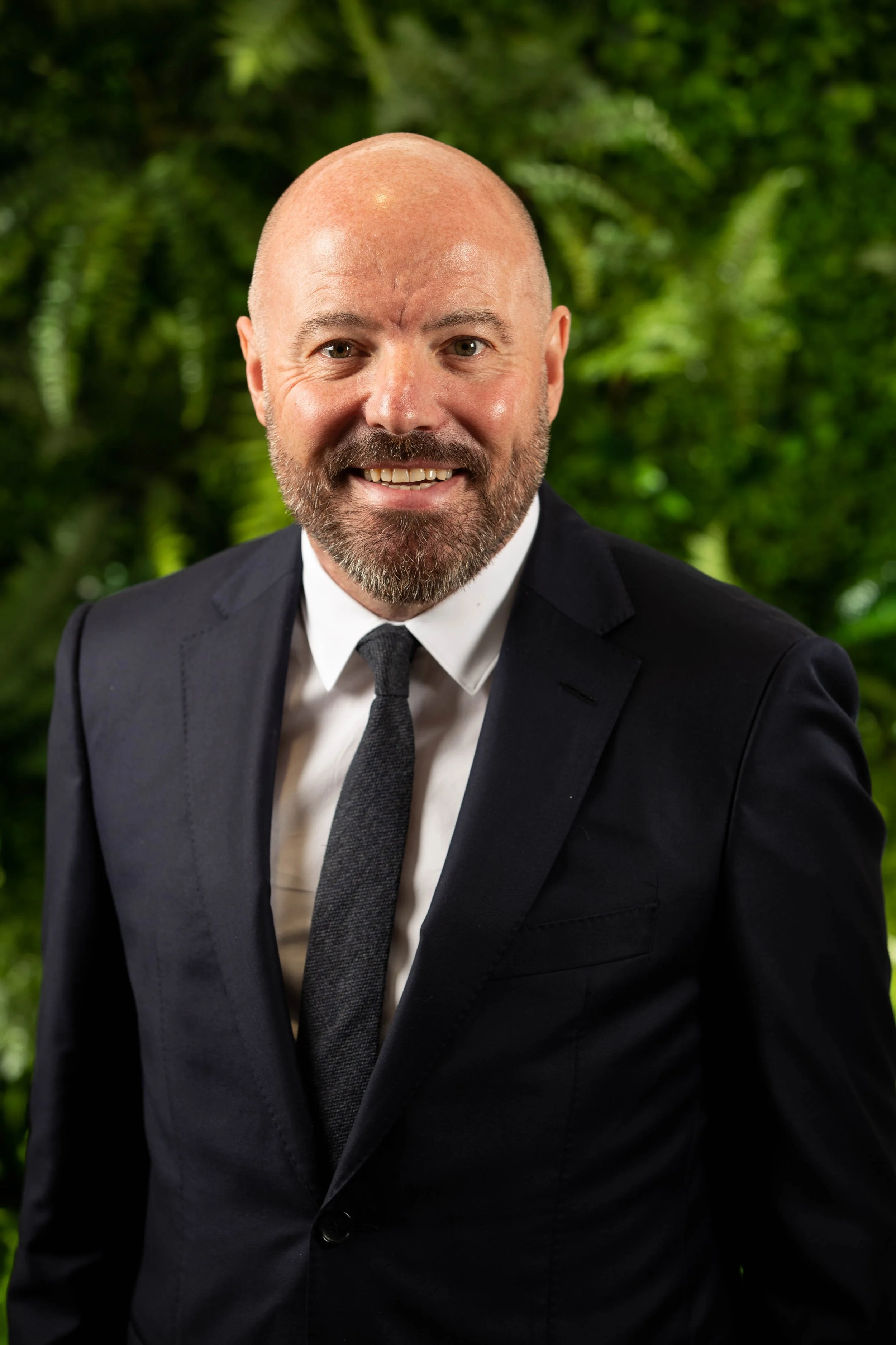 A smiling man with a beard and bald head, wearing a gray checkered blazer and light gray dress shirt, standing in front of a wall with framed pictures.