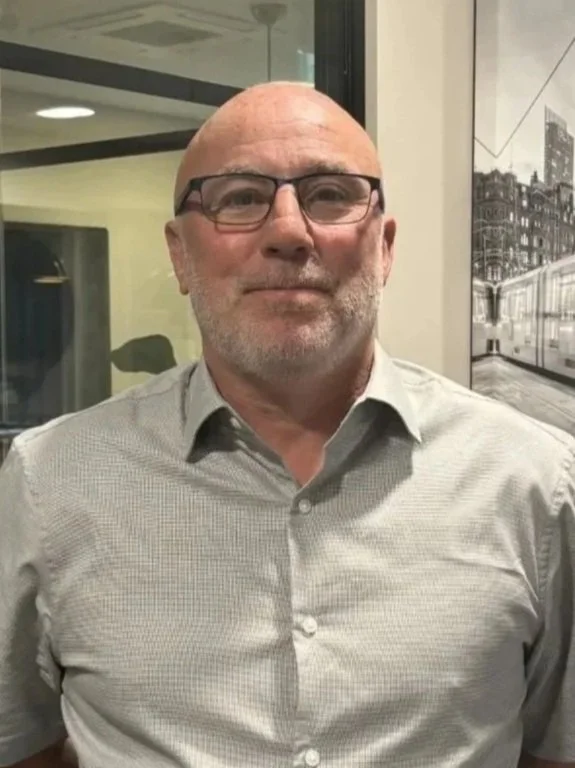 A smiling middle-aged man with gray hair and a beard, wearing a suit jacket and striped shirt, standing in front of framed artwork on a wall.