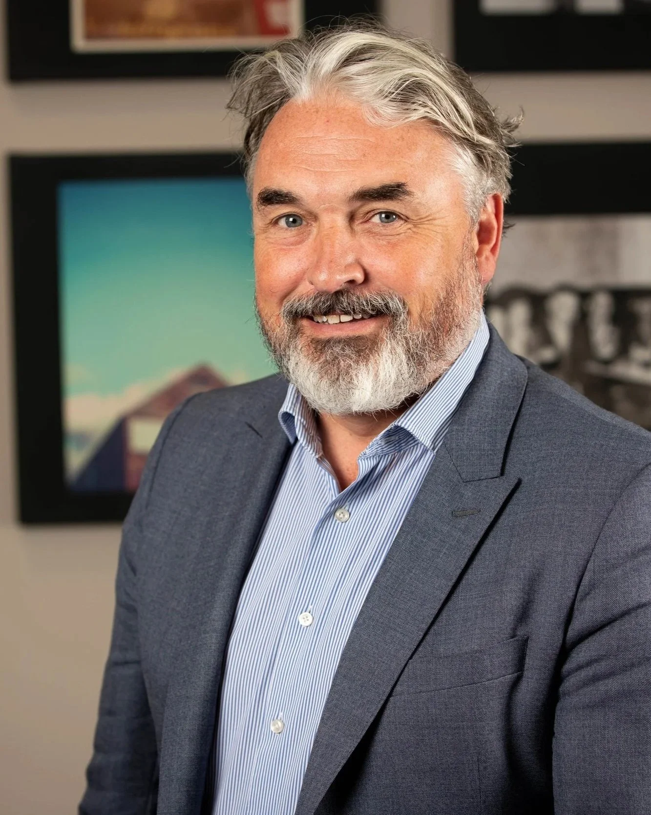 A smiling man with a beard and bald head, wearing a gray checkered blazer and light gray dress shirt, standing in front of a wall with framed pictures.