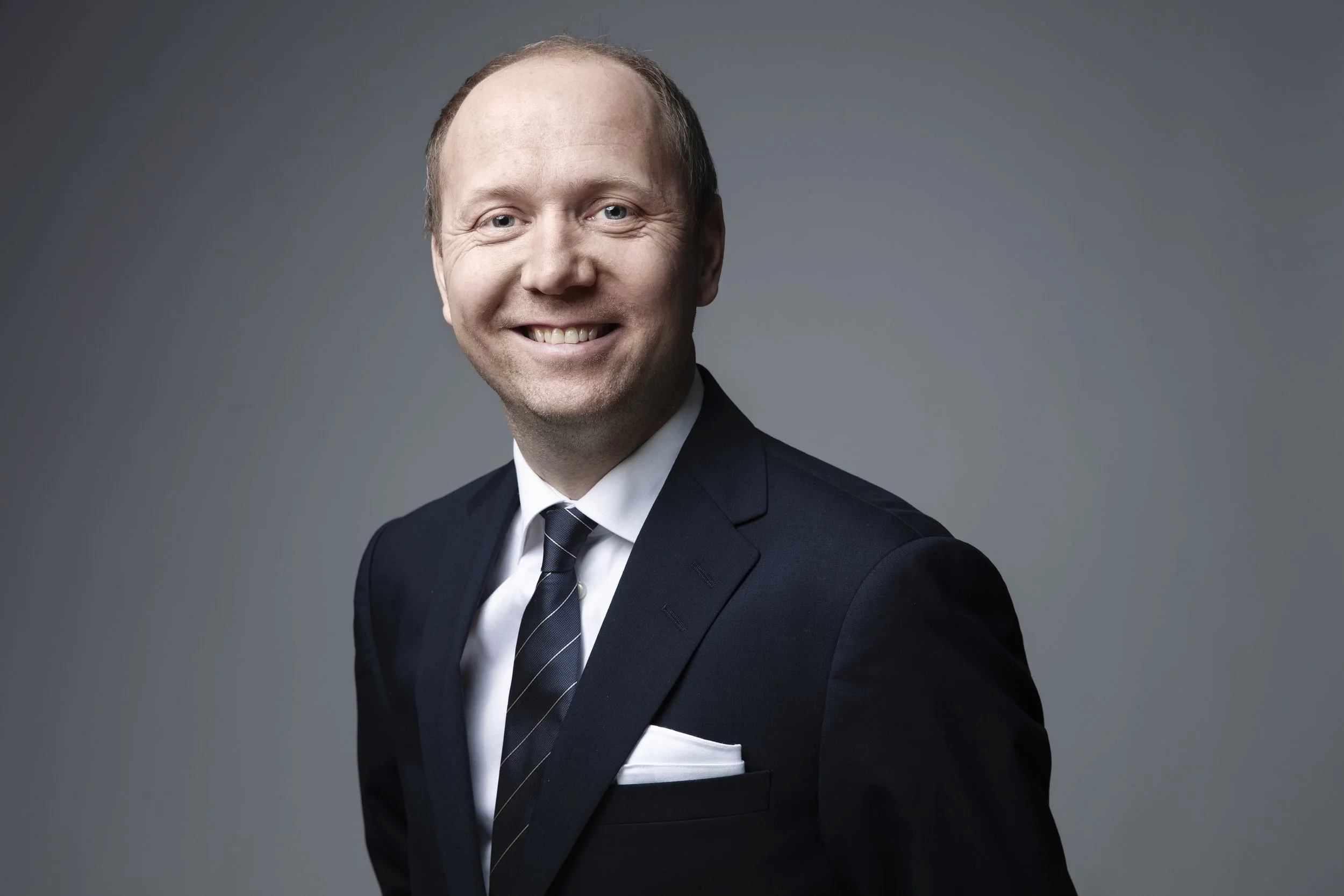 Et portrett av en mann i svart dress med hvit skjorte og stripete slips, smilende mot kamera mot en grå bakgrunn. Thomas Ryste