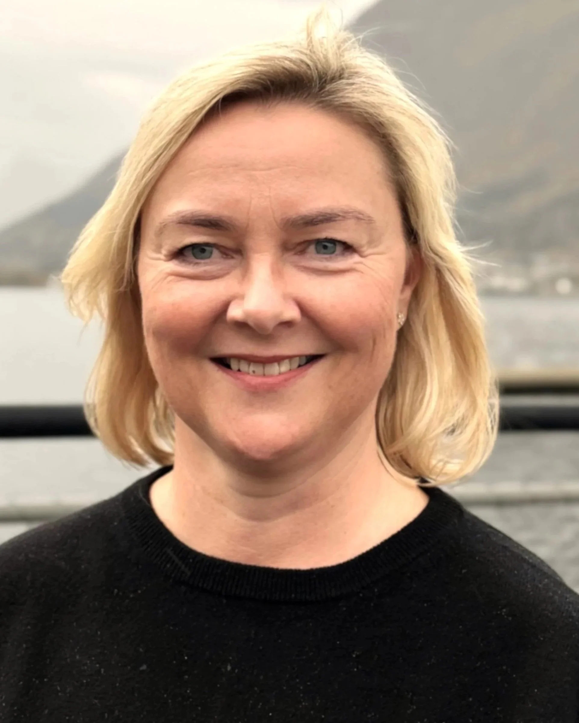 Et portrett av en smilende kvinne med blond hår, iført svart genser, med fjordlandskap i bakgrunnen.
