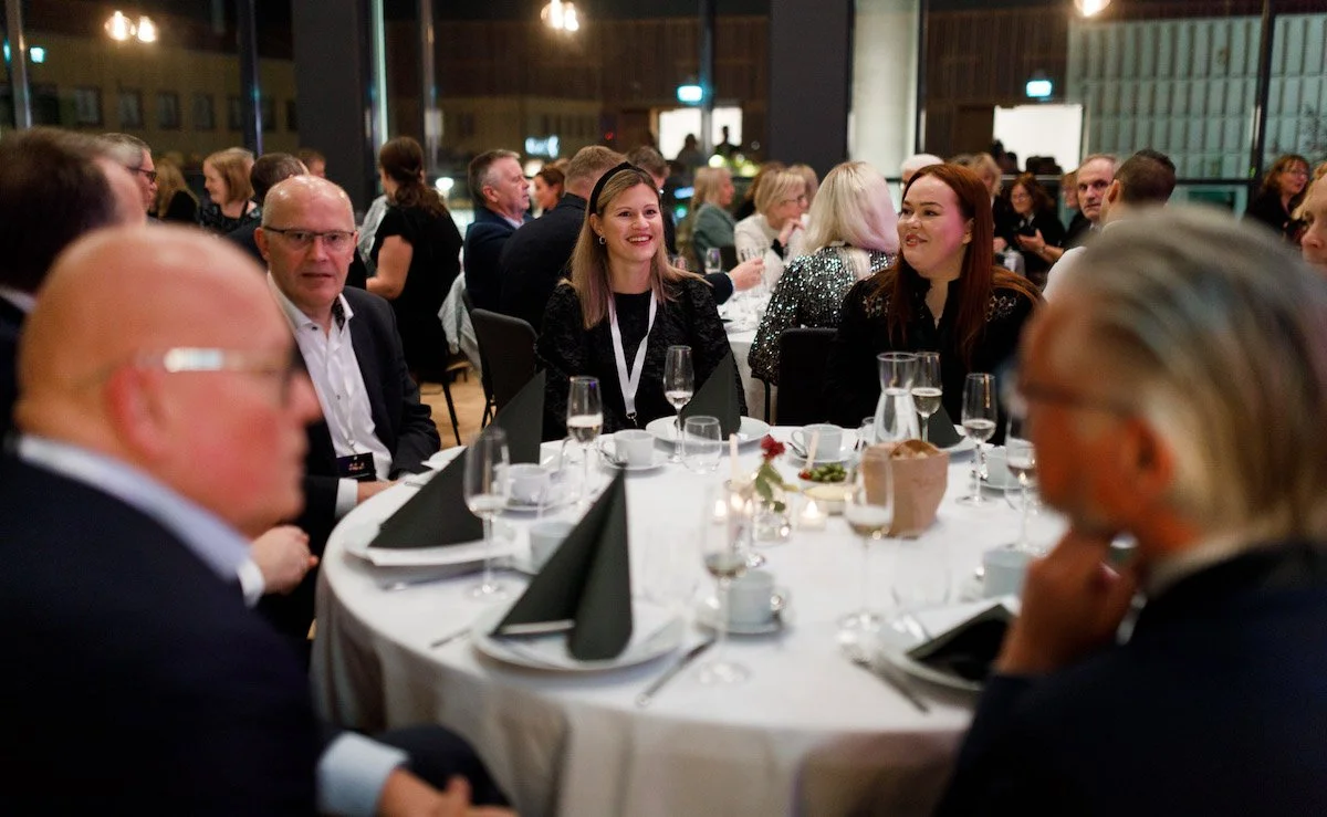 Mennesker som spiser og snakker ved et rundt bord under en formell sammenkomst. Innendørs med store vinduer og bylys utenfor.