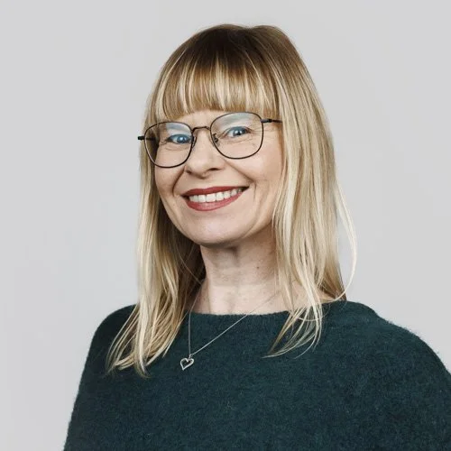 Portrett av en smilende kvinne med blond hår, briller og et mørkt antrekk