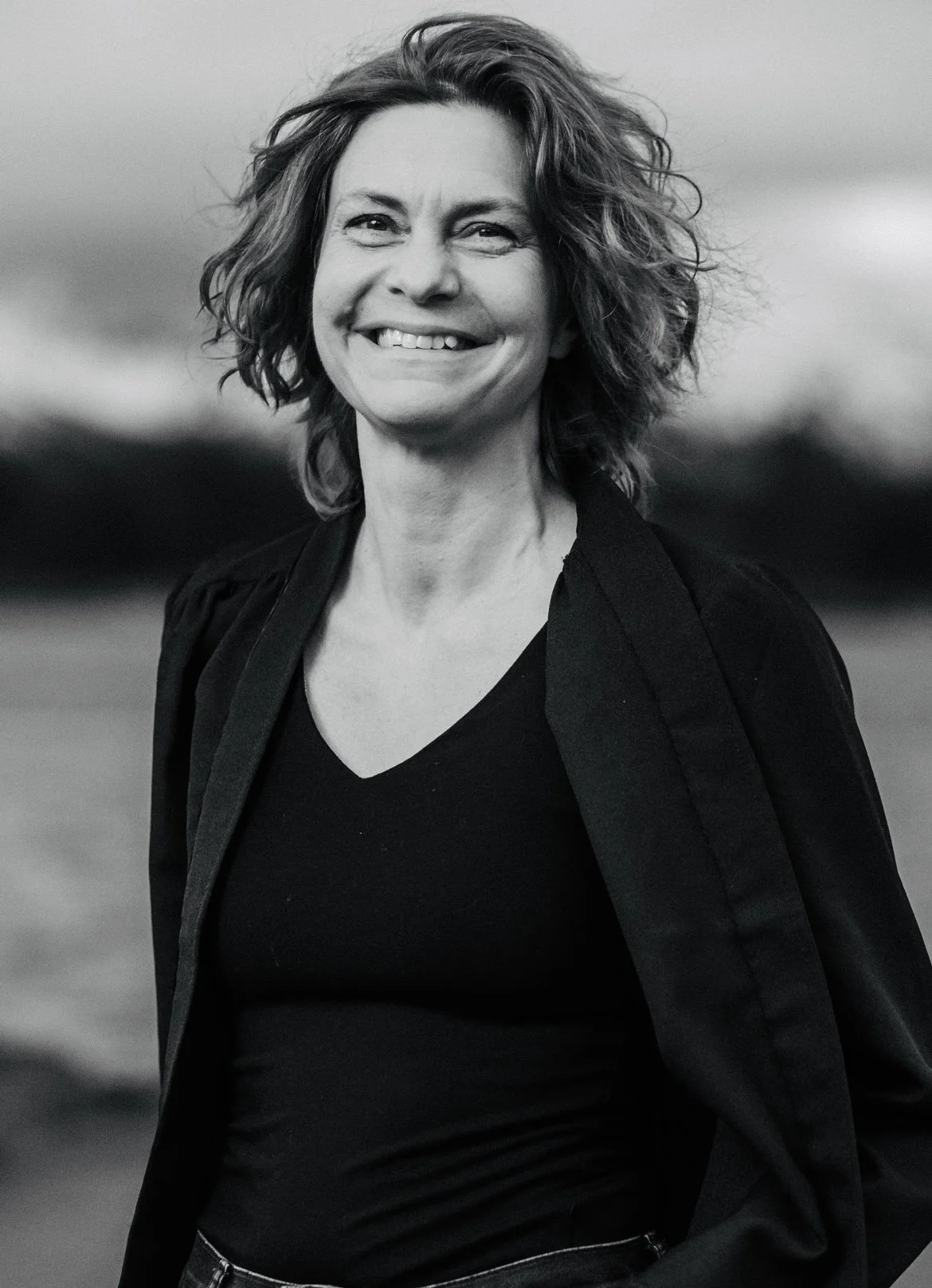 Et svart-hvitt bilde av en smilende kvinne med krøllete hår, iført svart T-skjorte og svart jakke, utendørs med uklart landskap i bakgrunnen.