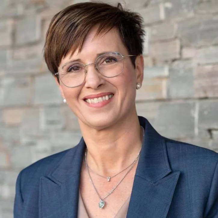 En kvinne med kort brunt hår, iført briller og formell blazé, smilende foran en steinvegg.