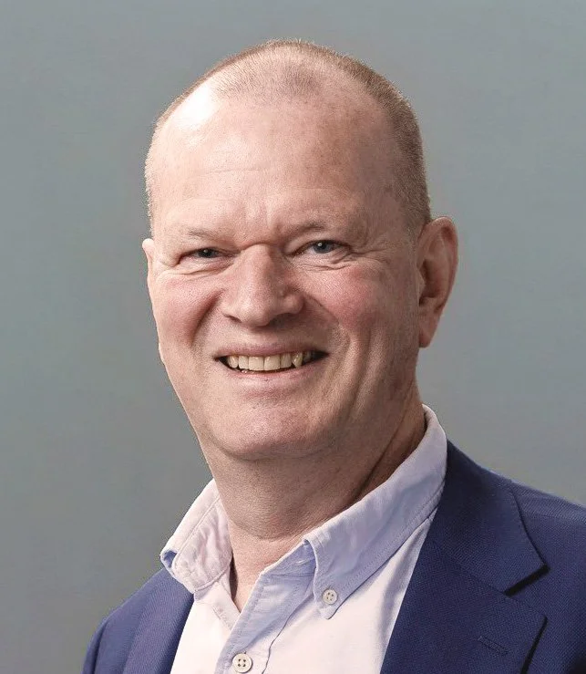 Portrett av en mann med lyst hår, blå dress og hvit skjorte, smilende mot kameraet.