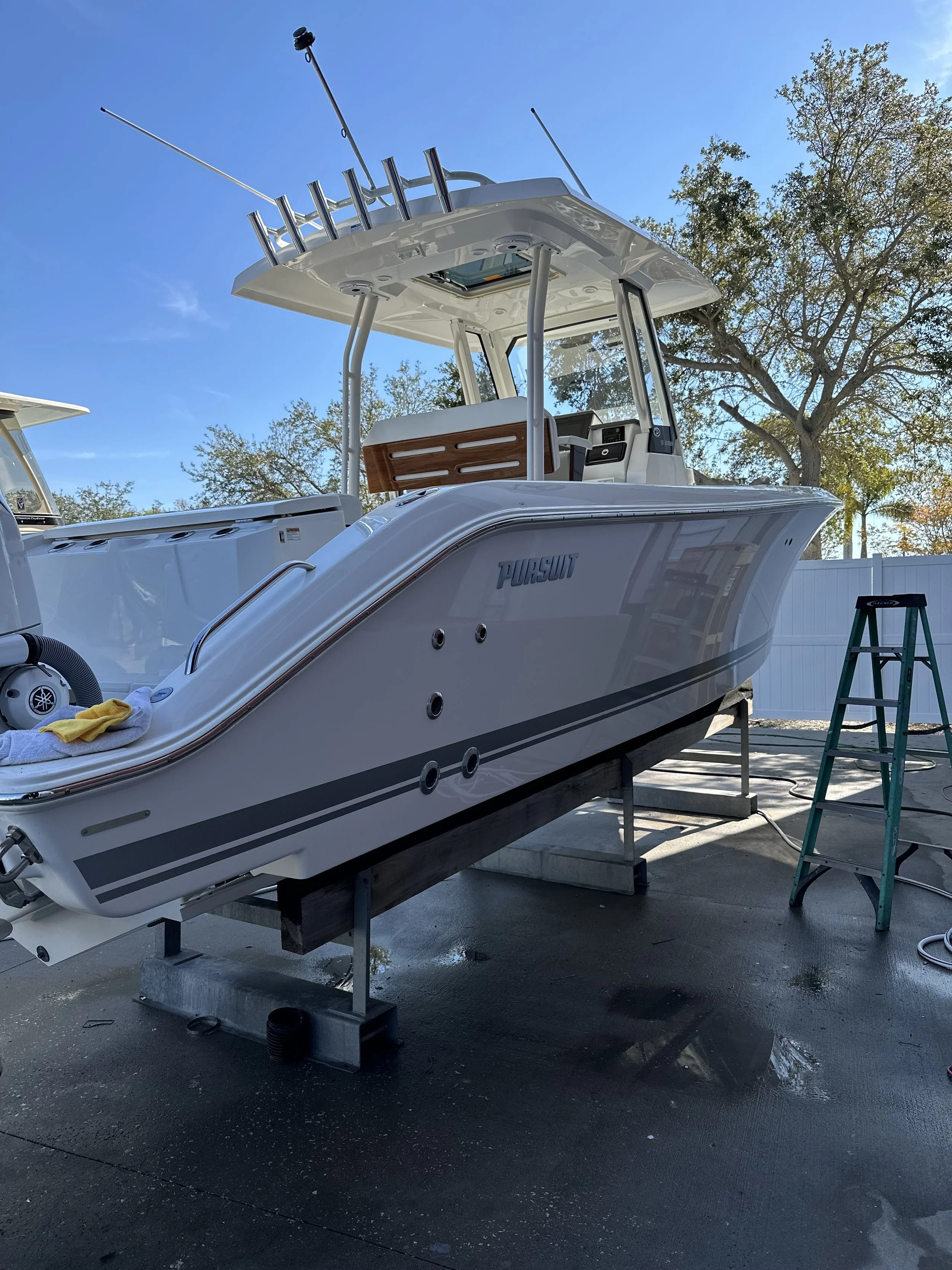 A white Pursuit boat on a trailer, outdoors, with a clear blue sky, trees, and a white fence in the background.