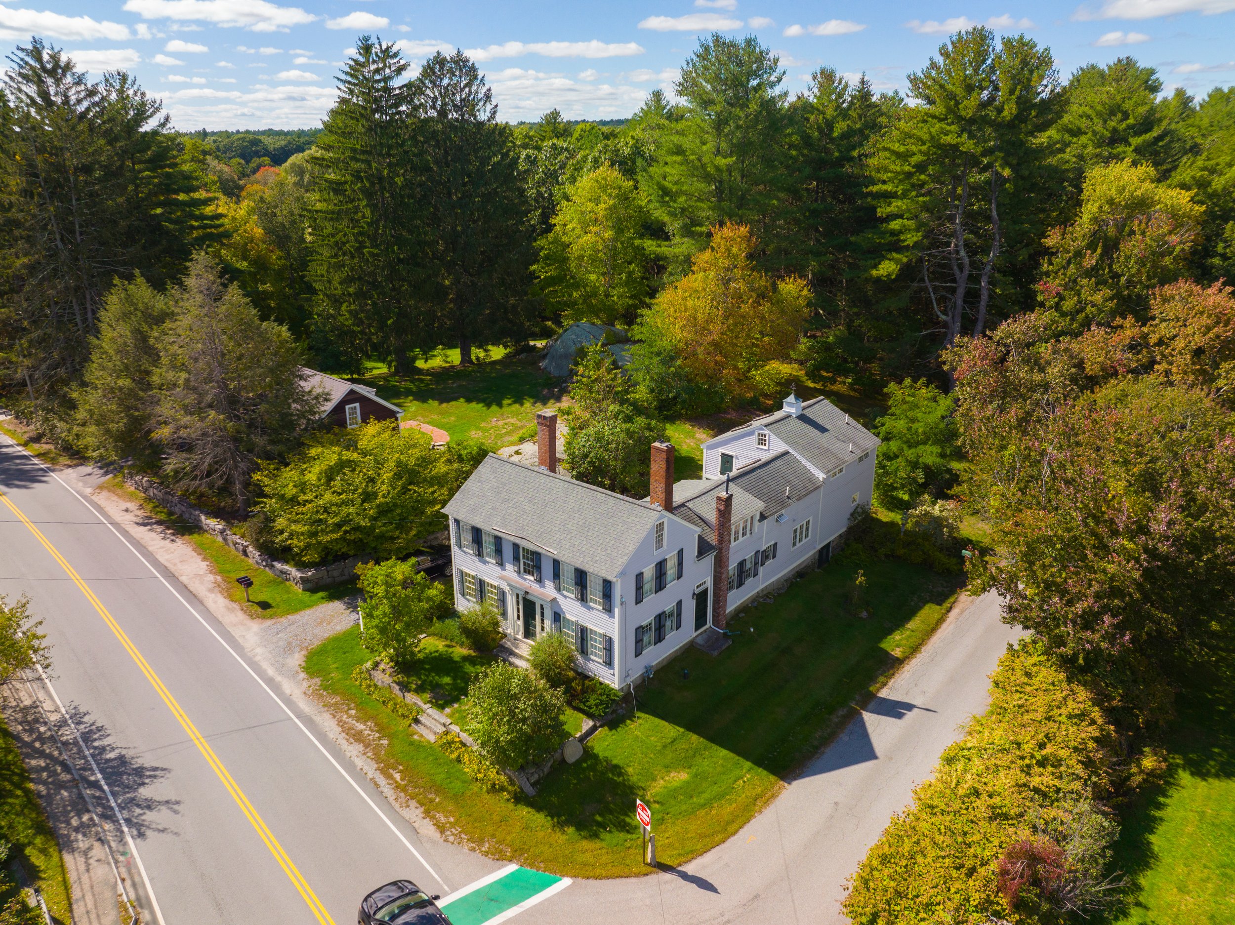 Aerial view of a large white colonial-style house surrounded by trees and gardens, positioned at the corner of an intersection on a suburban street. The house has multiple chimneys and a well-maintained lawn.