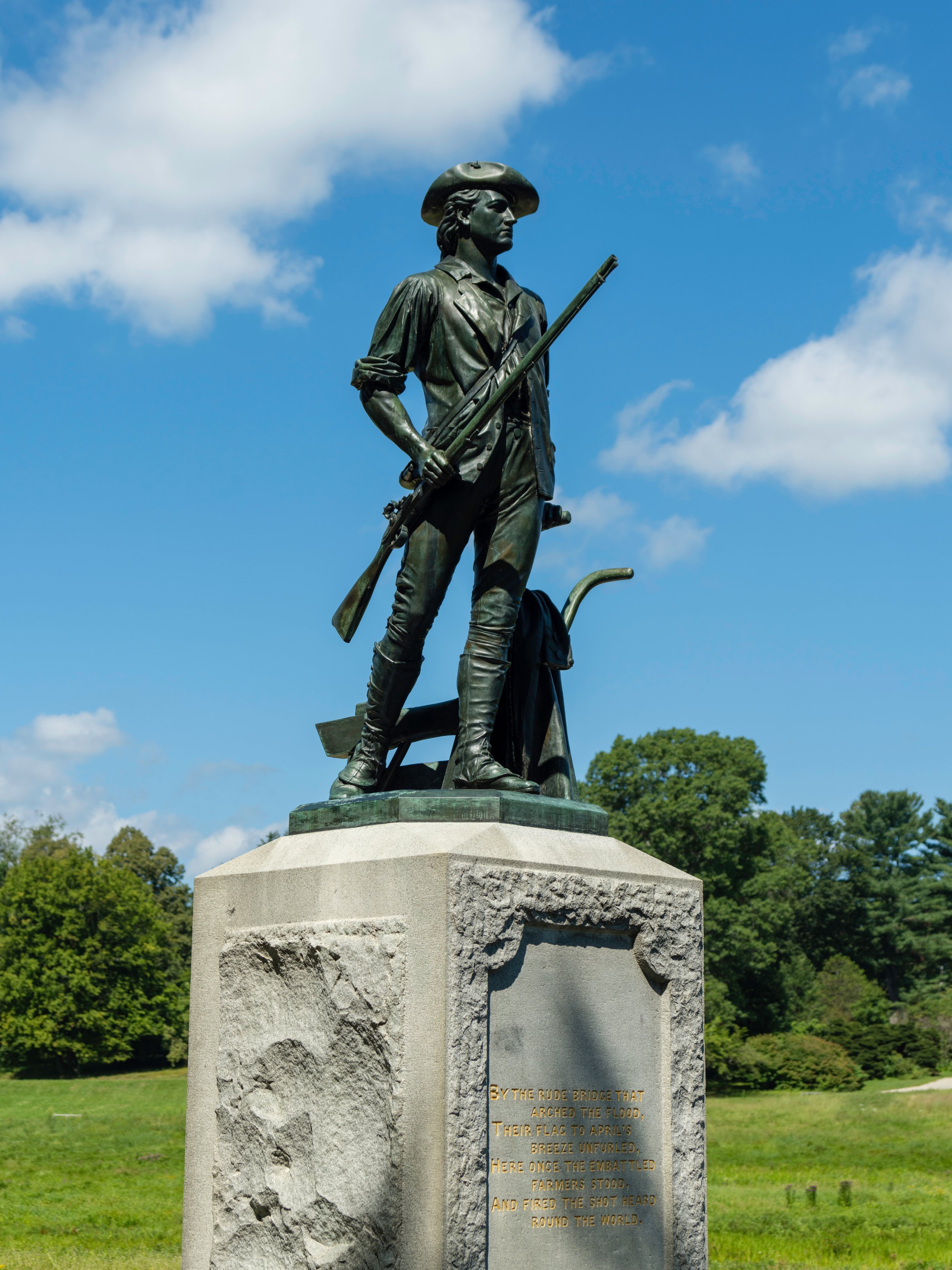 Statue of a Revolutionary War soldier holding a rifle on a pedestal, set in a grassy landscape with trees and a blue sky with clouds.