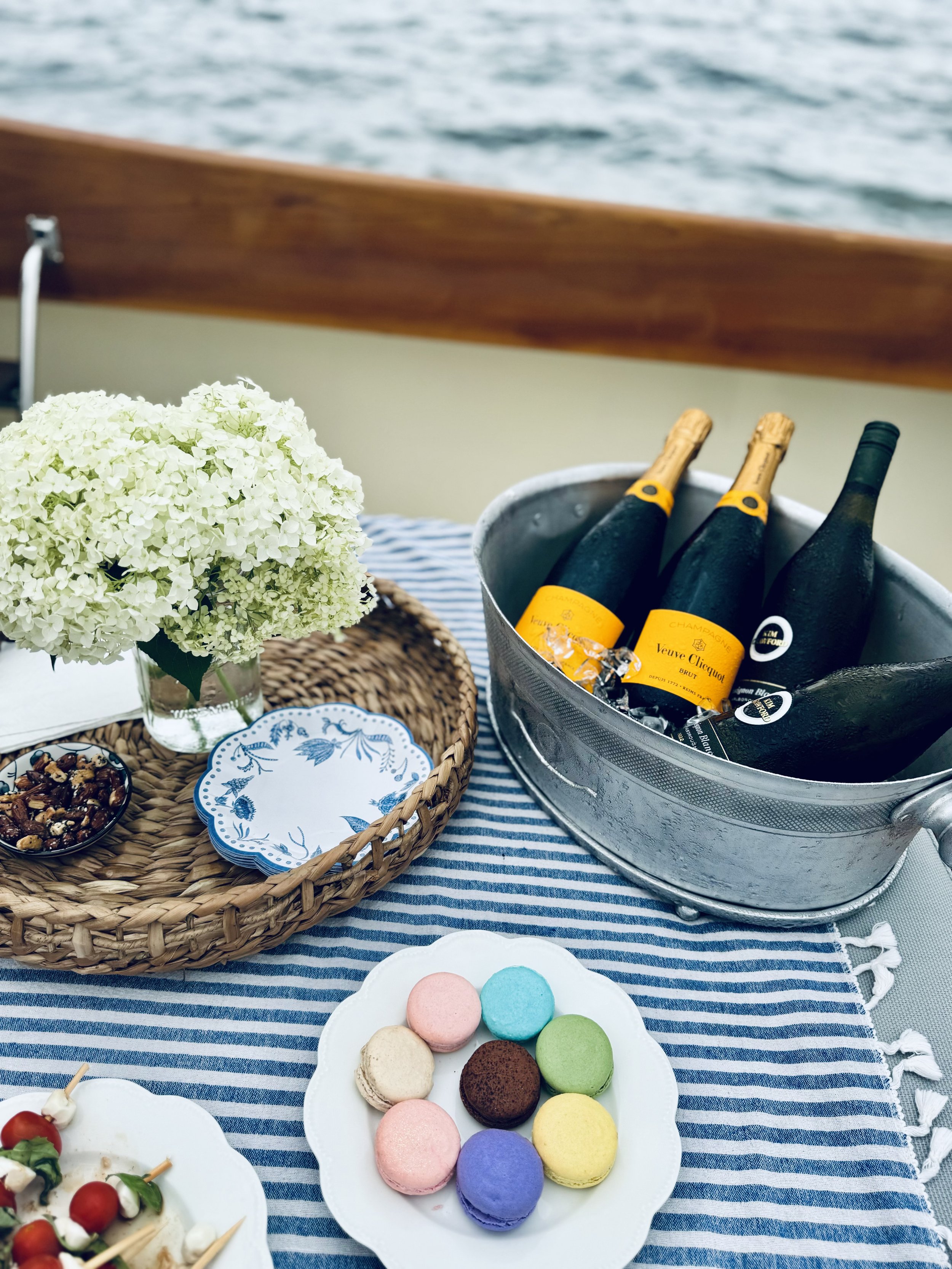 A table set on a boat with a bouquet of white flowers in a vase, a wicker tray with patterned napkins, a bowl of mixed nuts, a plate of colorful macarons, skewers with tomatoes and mozzarella, and a metal bucket containing bottles of champagne agains