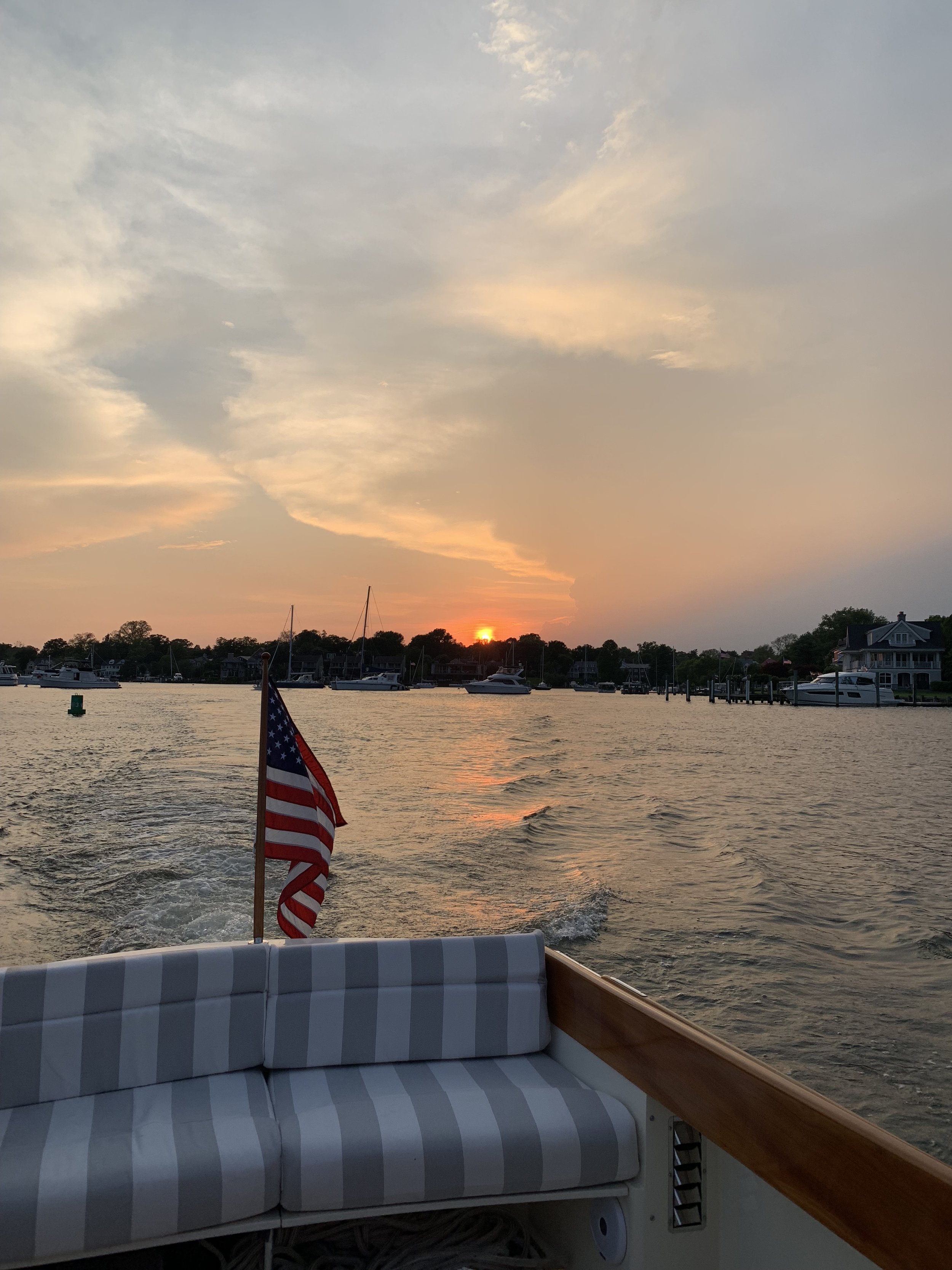 Sunset view from a boat with American flag, striped seating, calm water, distant boats, and shoreline.