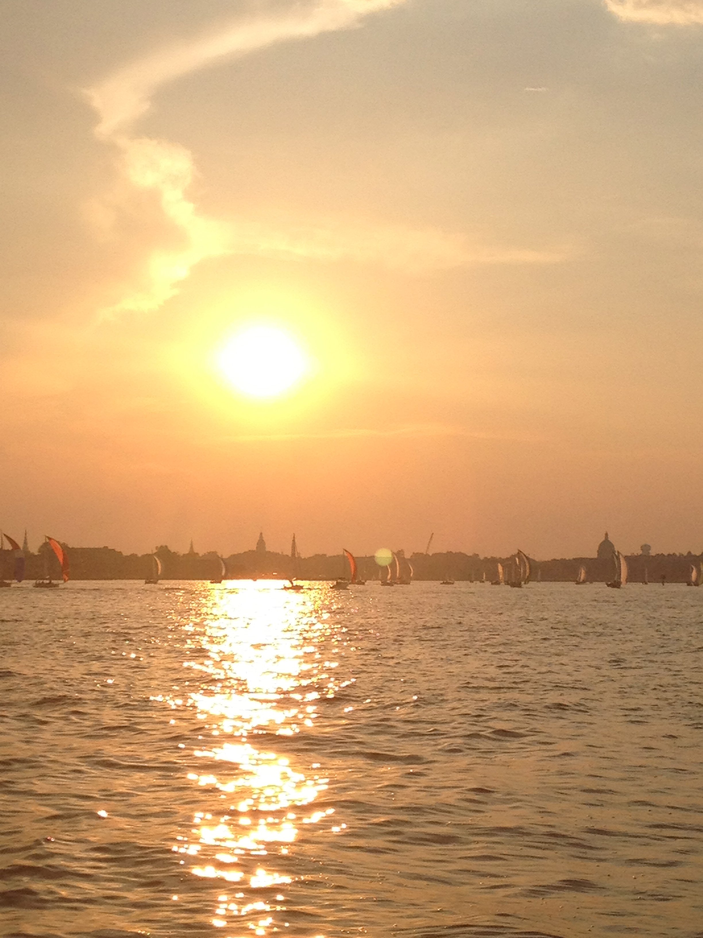 Sunset over water with sailboats on the horizon, reflecting golden light on the surface, and a clear sky with few clouds.
