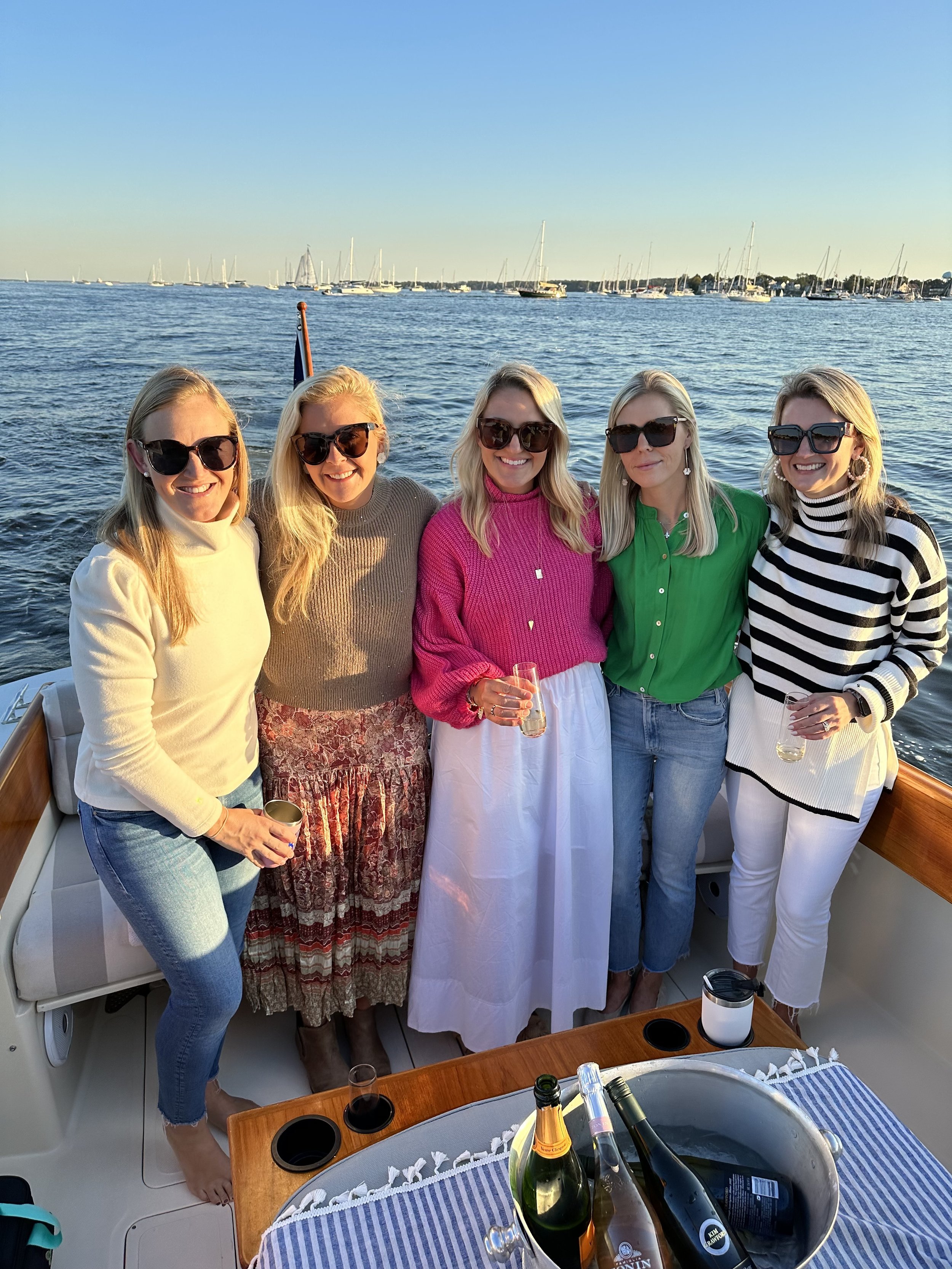 Five women on a boat holding drinks with a scenic ocean background and sailboats in the distance.