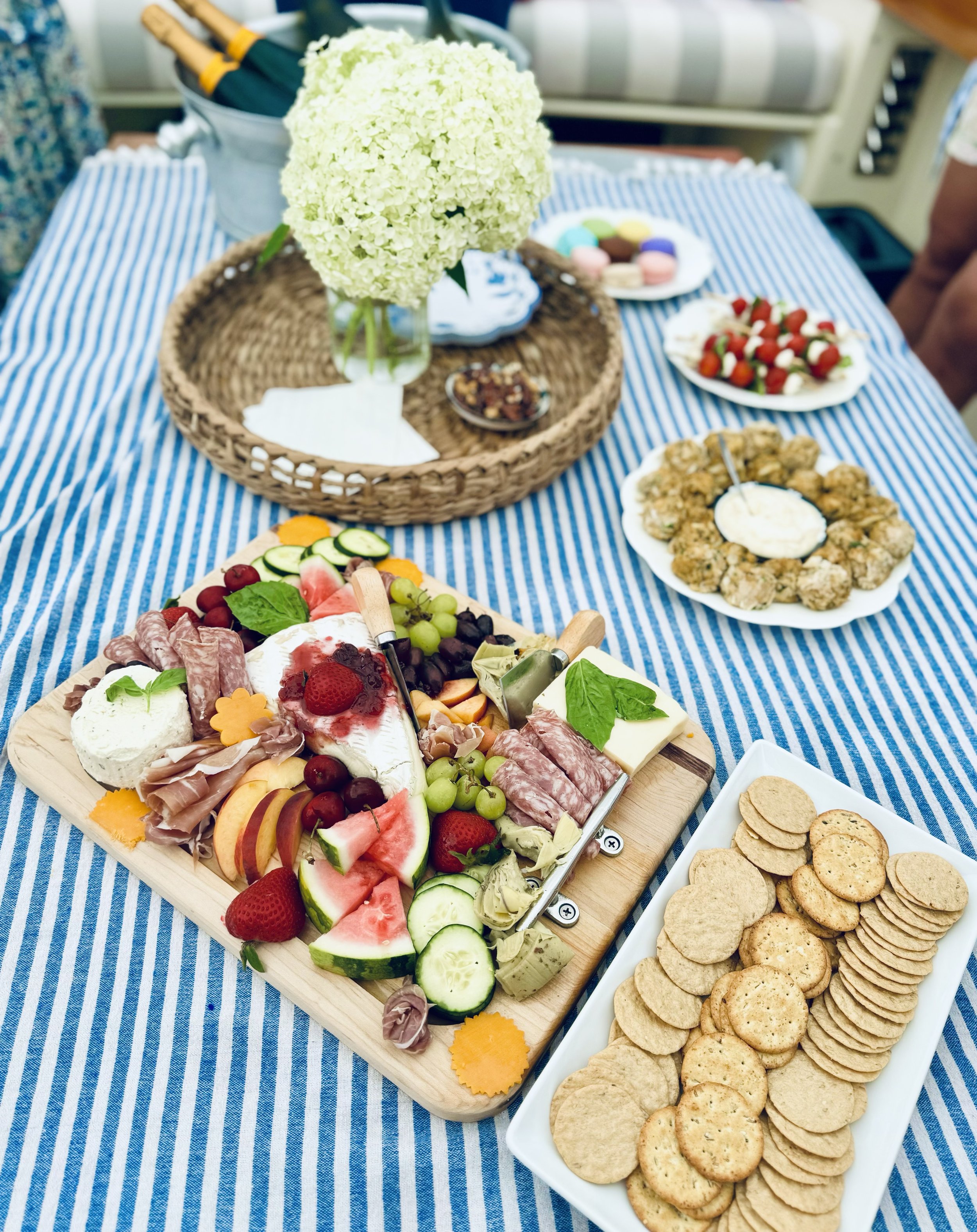 A charcuterie board with assorted cheeses, meats, fruits, and vegetables on a blue and white striped tablecloth. Nearby are plates of crackers, macarons, and appetizer bites, with a bouquet of white flowers in a basket and two bottles in a bucket.