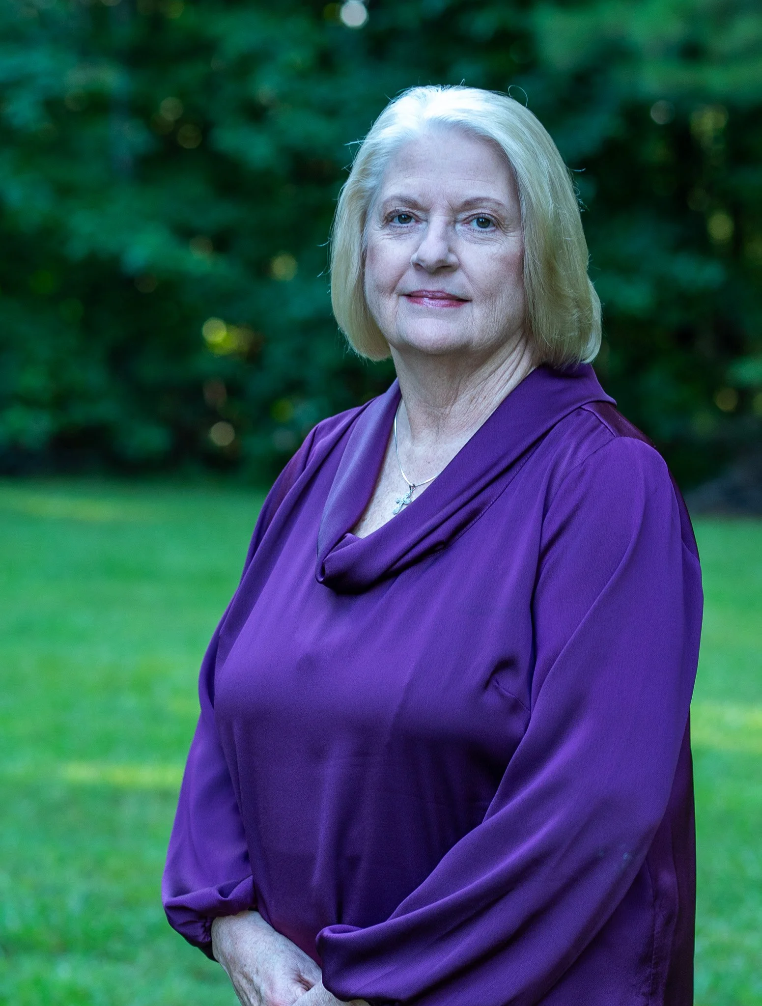 An older woman with blonde hair wearing a purple dress standing outdoors in front of green trees and grass.