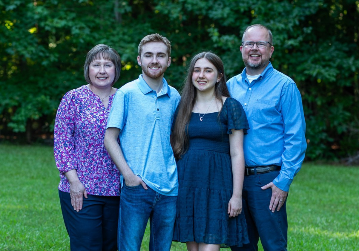 Family of four standing outdoors on a grassy area with green trees in the background, smiling at the camera.