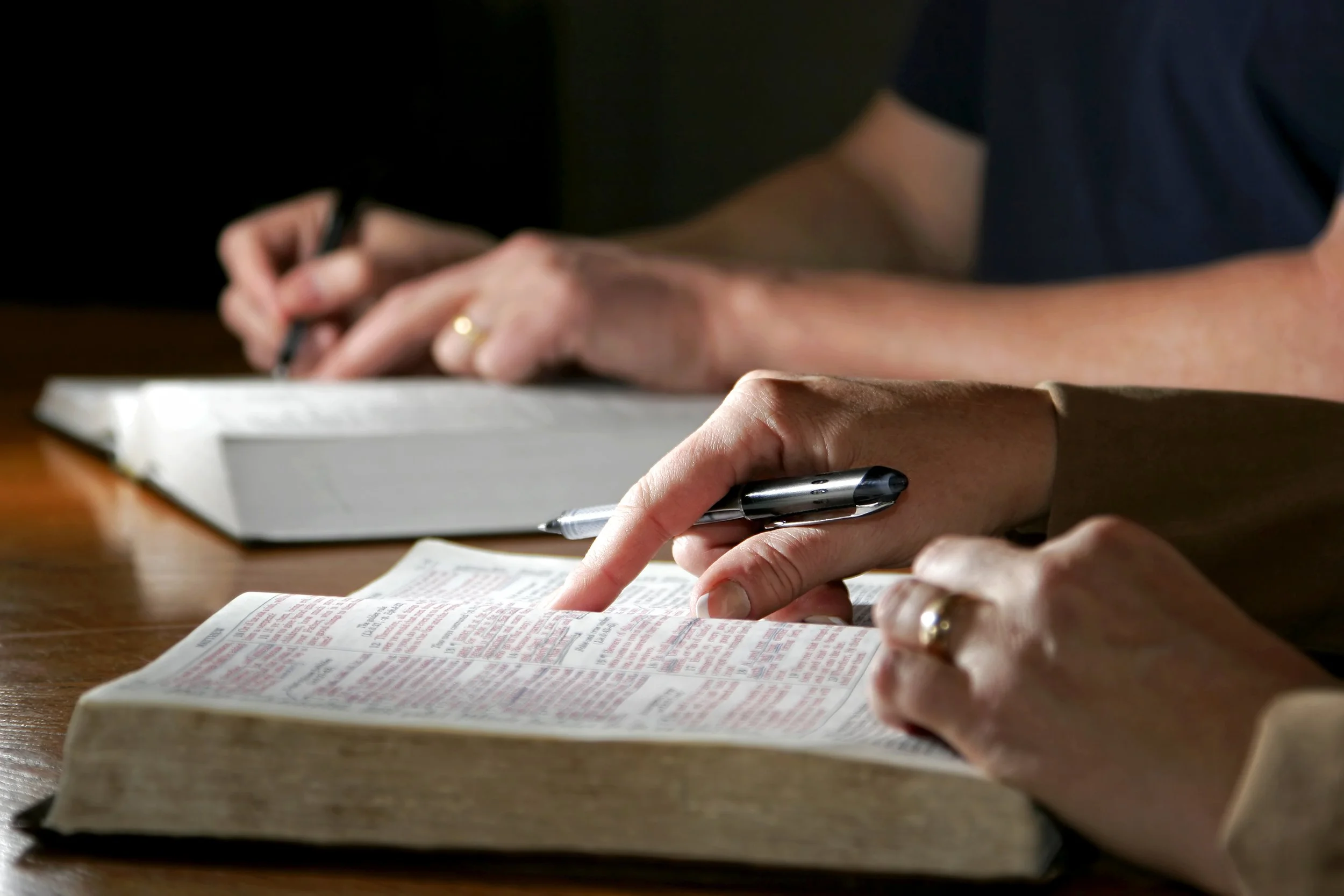 Two people reading or studying a Bible at a wooden table, with one person pointing to a verse and the other holding a pen.