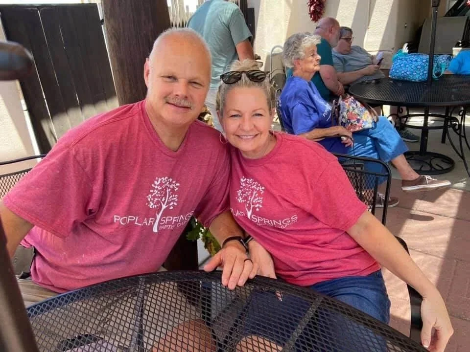 A smiling man and woman sitting together at an outdoor table, wearing matching pink t-shirts with a tree logo and the words "Spring Poplar" on them, with older women in the background.
