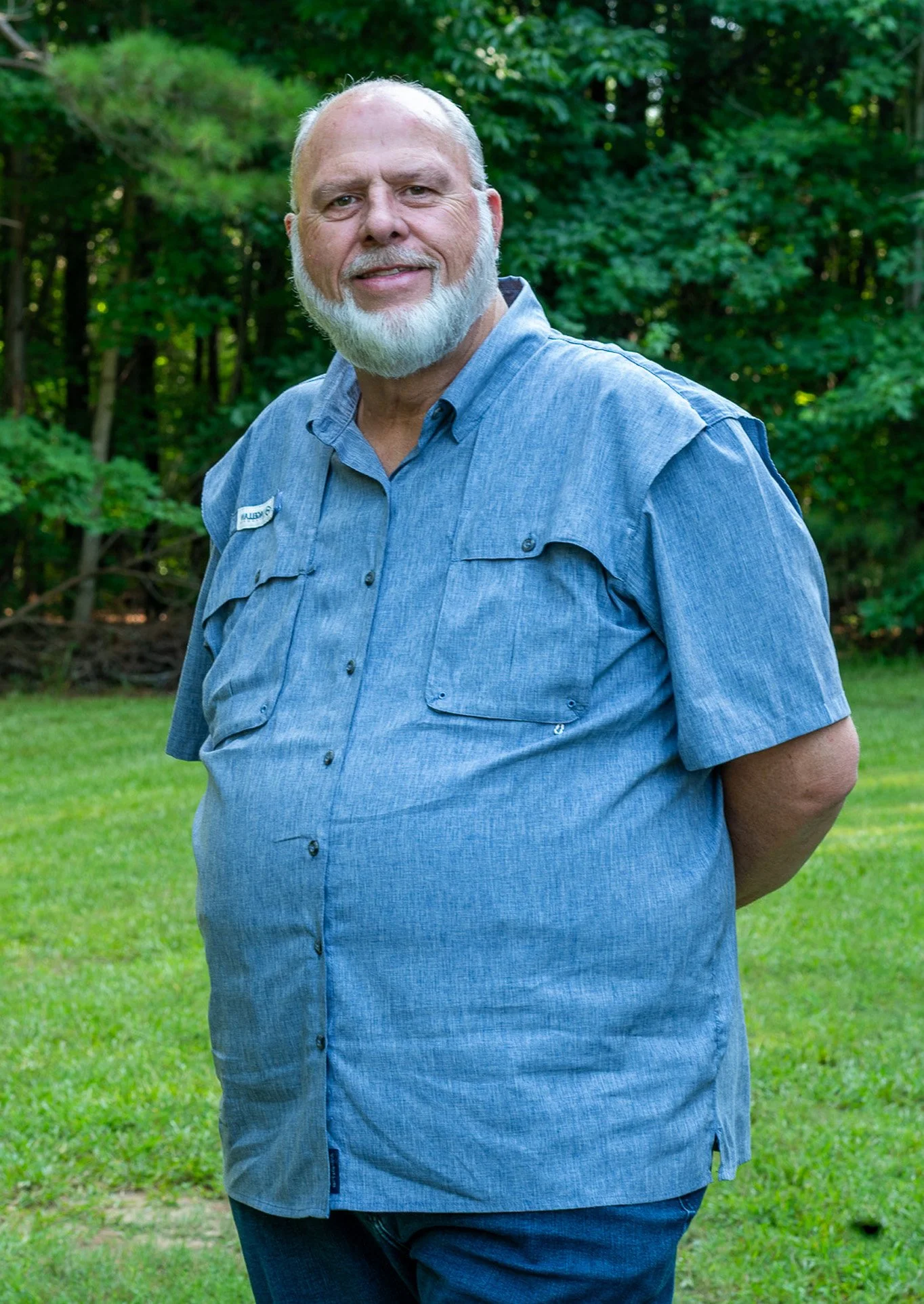 An older man with a white beard and short hair standing outdoors on a grassy area, wearing a blue short-sleeved button-up shirt and smiling.