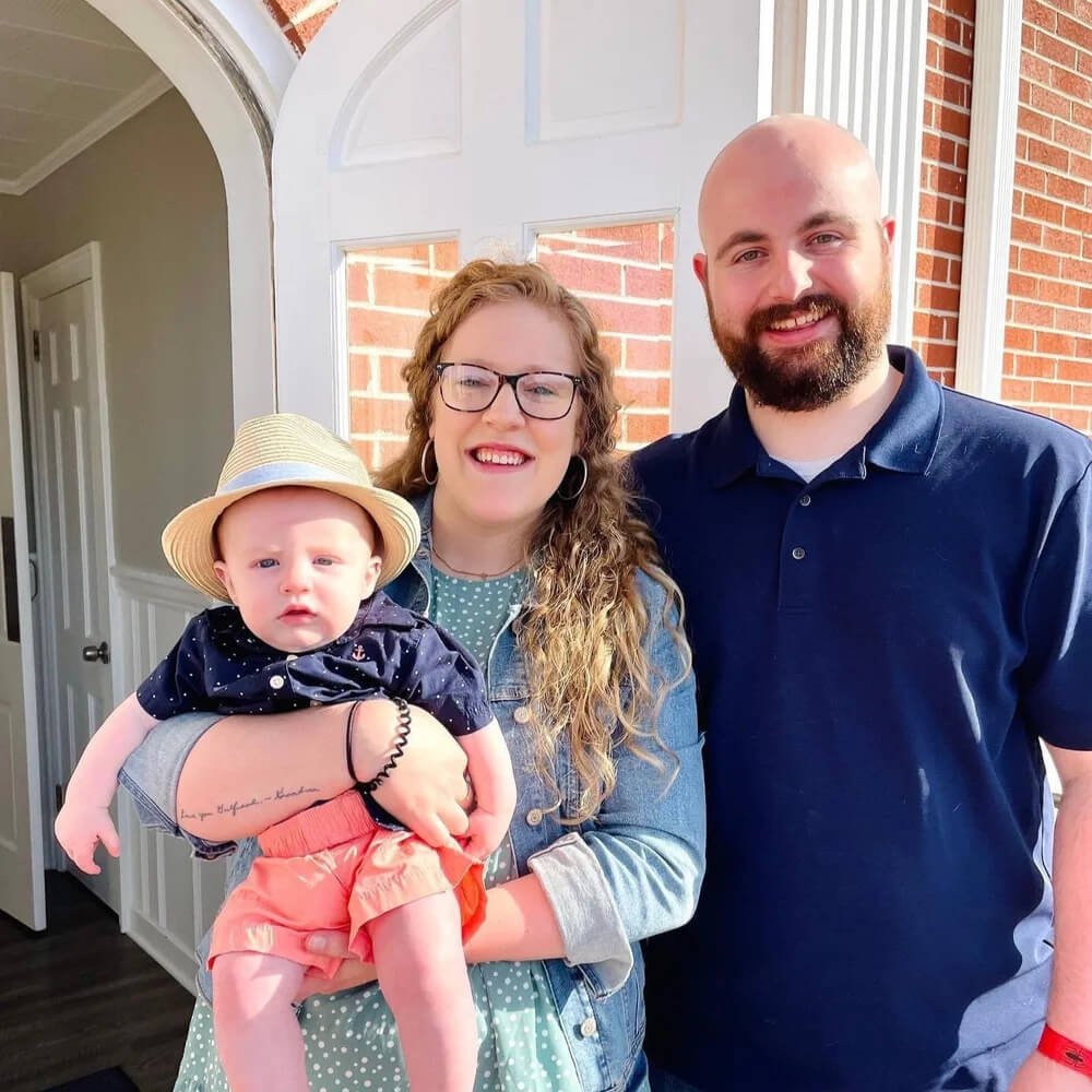 A smiling family of three standing outdoors in front of a brick house. The woman is holding a baby boy, and there is a man standing next to her.