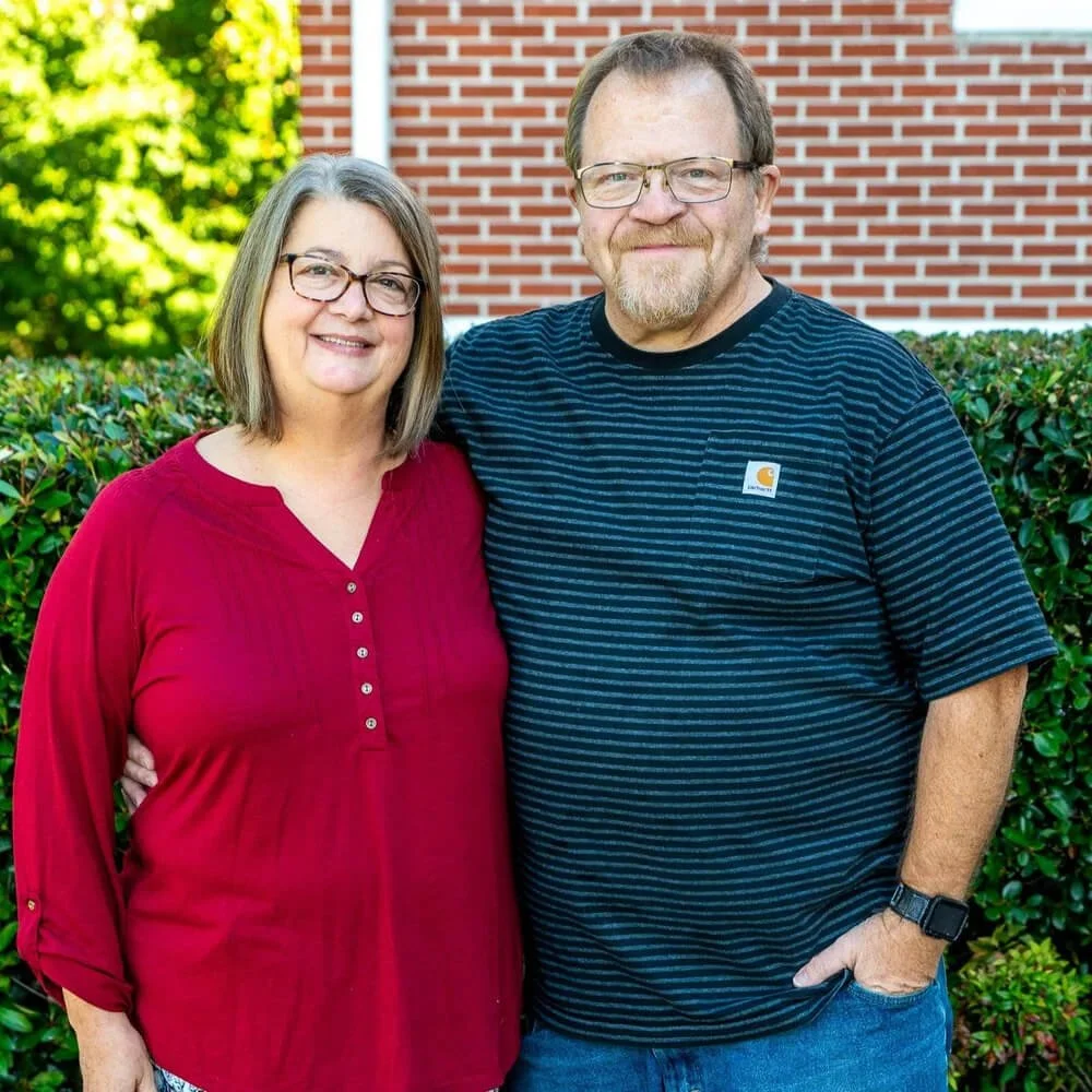A smiling woman and man standing outdoors in front of a green hedge and a brick building, with sunlight filtering through trees in the background.