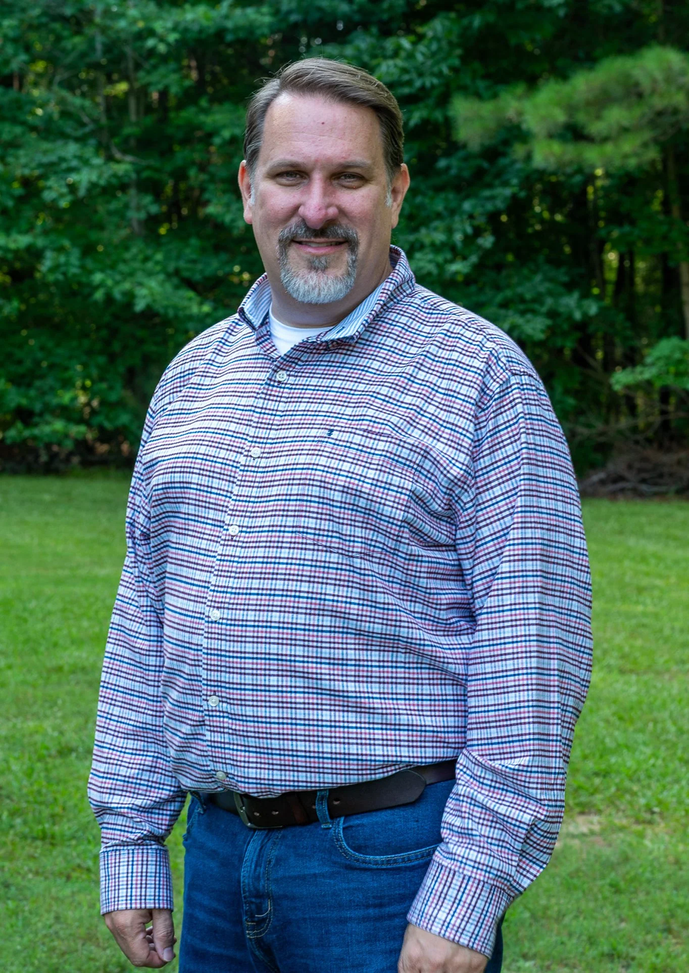 A man with short brown hair, a beard, and mustache standing outdoors on a grassy area with trees in the background, wearing a checked button-up shirt and blue jeans.