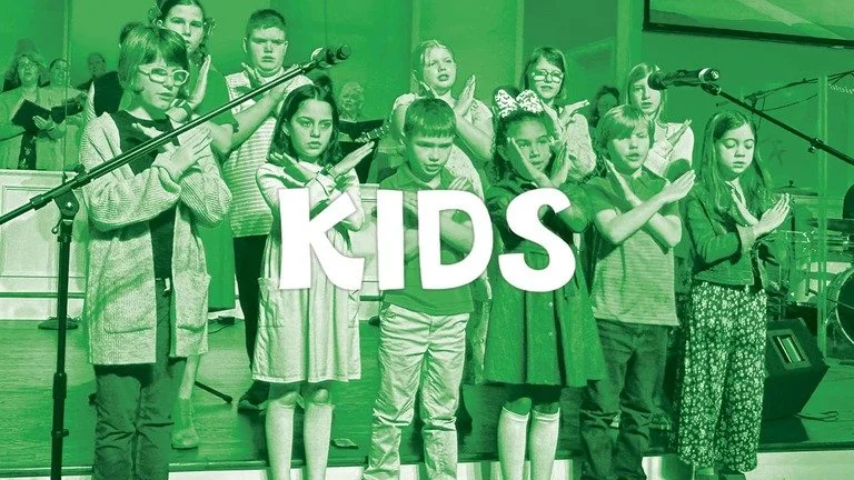 Children performing during a kids' event on stage, with some holding microphones, and a large sign reading 'KIDS' in front of them.