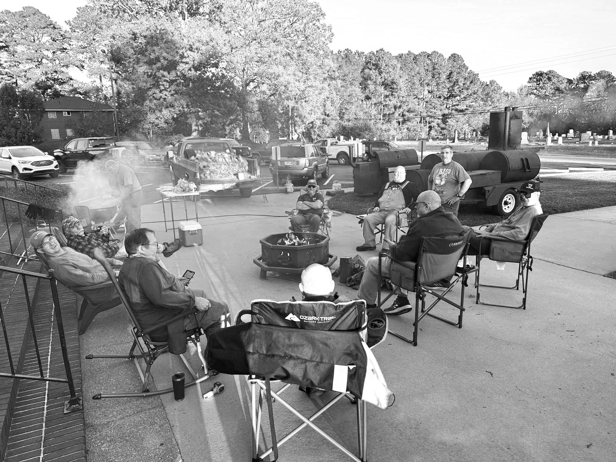 A group of people gathered outdoors around a fire pit is seen in this black-and-white photo. Some are sitting in chairs, others standing, with trucks, a grill, and a barbecue smoker in the background. The setting appears to be a casual outdoor gathering, possibly a barbecue or cookout.