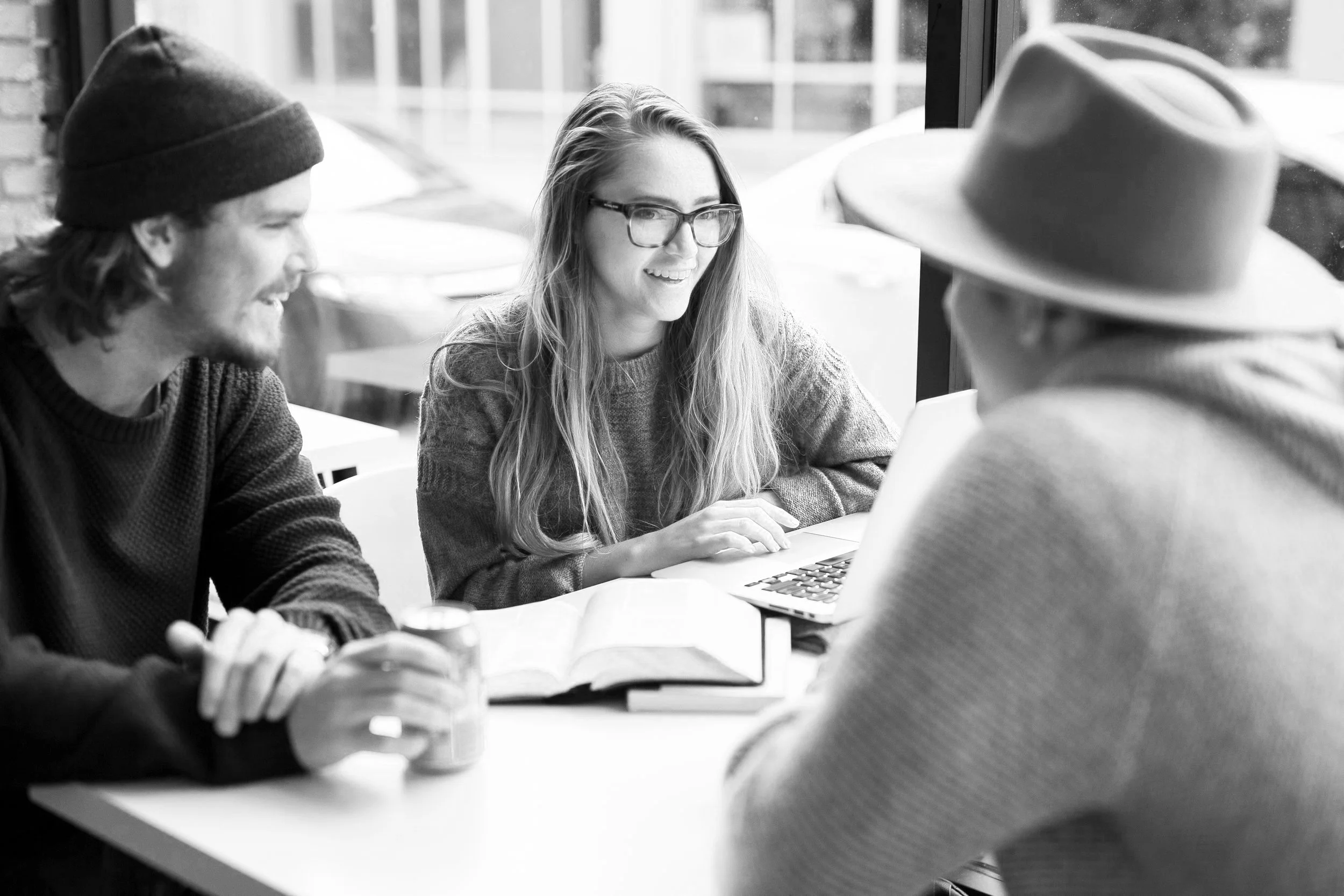 Three young adults sitting at a table, engaged in a conversation, with an open laptop and a notebook on the table, near a large window in a cafe or similar setting.