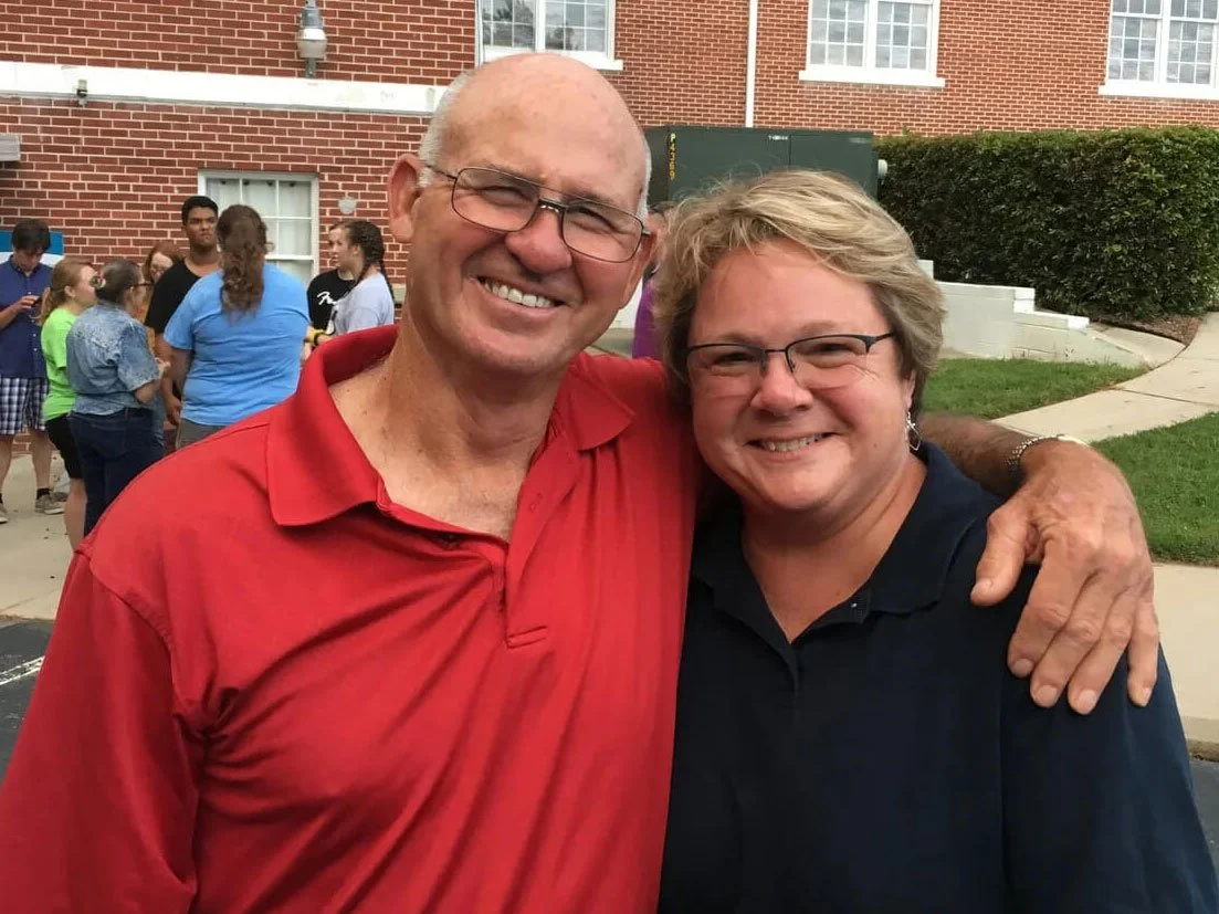 Two smiling men with glasses, one in a red polo shirt and the other in a black shirt, standing outside in front of a brick building with a group of people in the background.