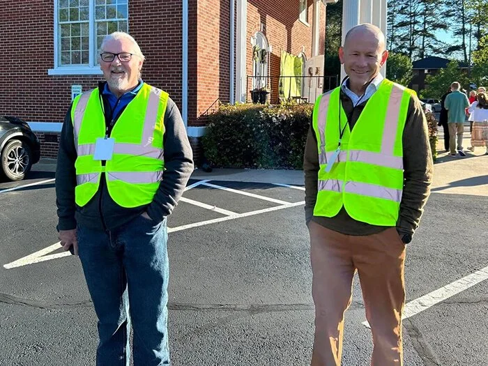 Two men wearing neon yellow safety vests standing outside in a parking lot near a brick building during daytime.